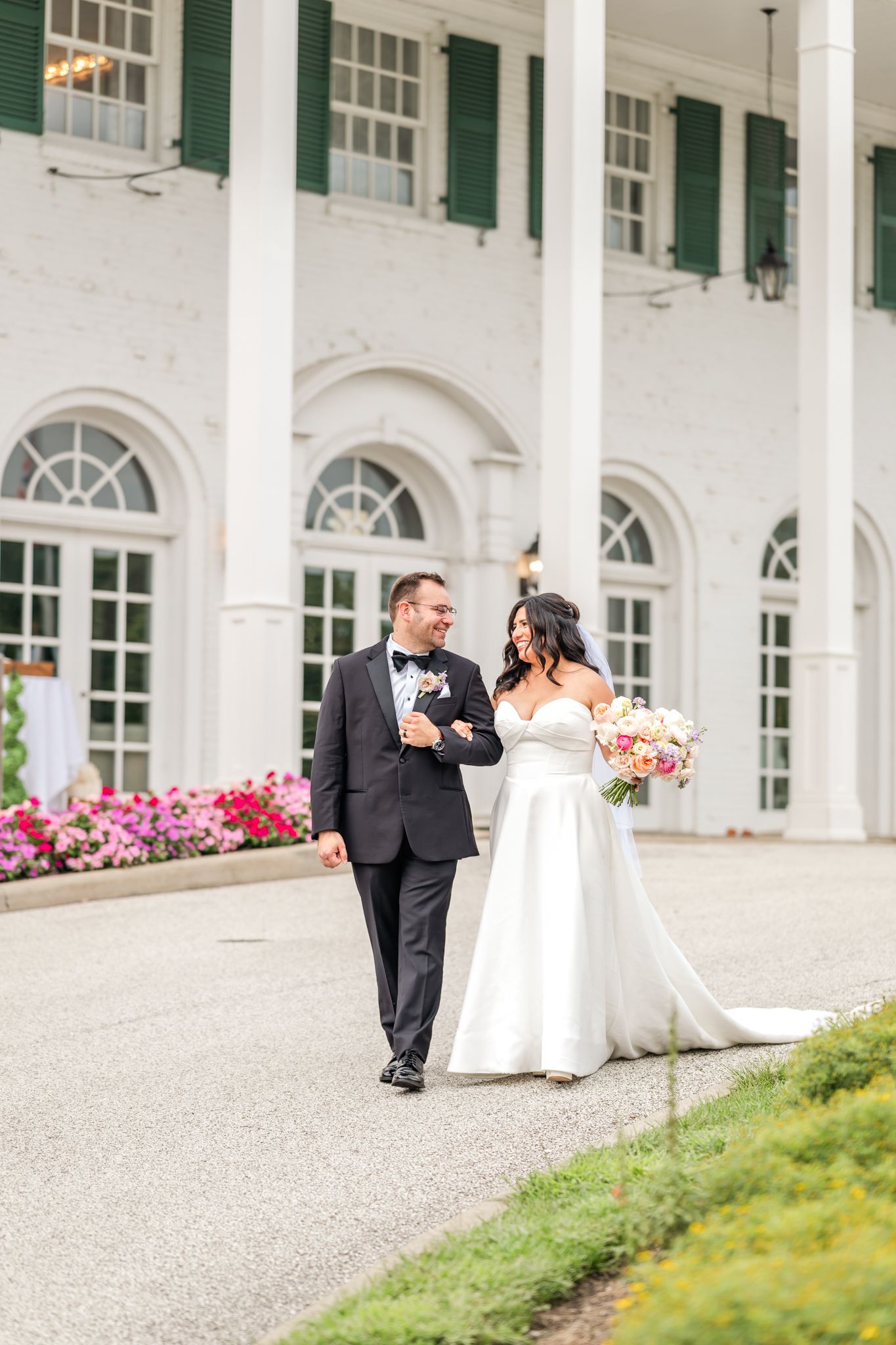 Bride in a white strapless gown and groom in a black tuxedo walk arm in arm past a white columned mansion with a pink floral bouquet.