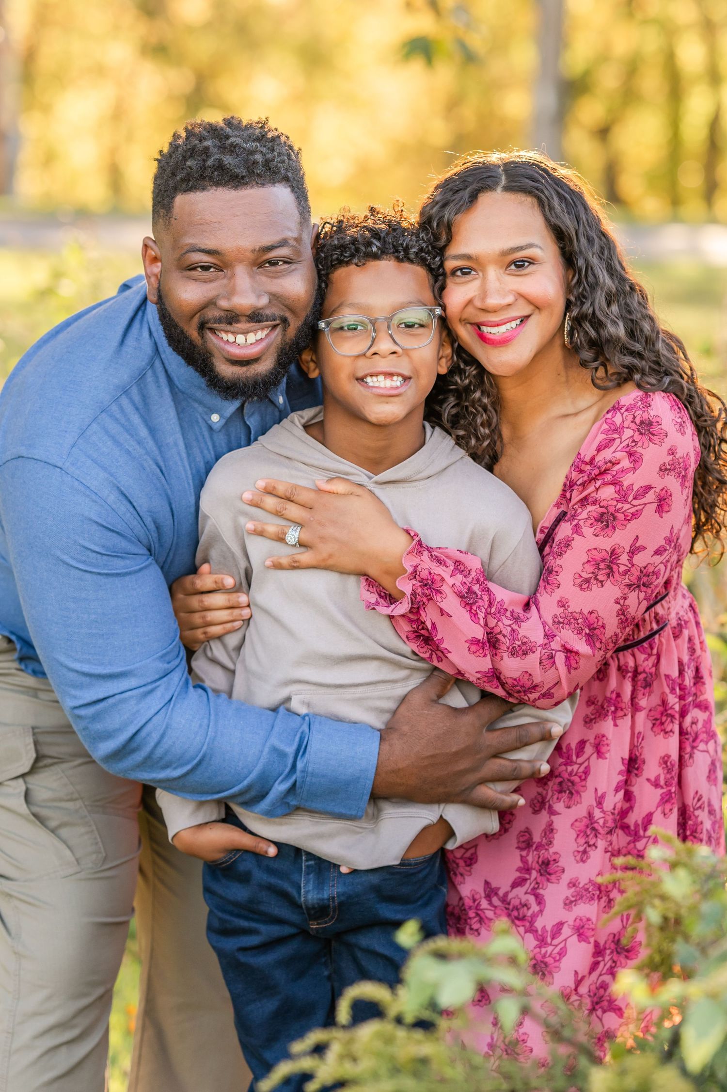 A smiling family of three embraces outdoors, with a boy in glasses between his bearded father and his mother in a pink floral dress.