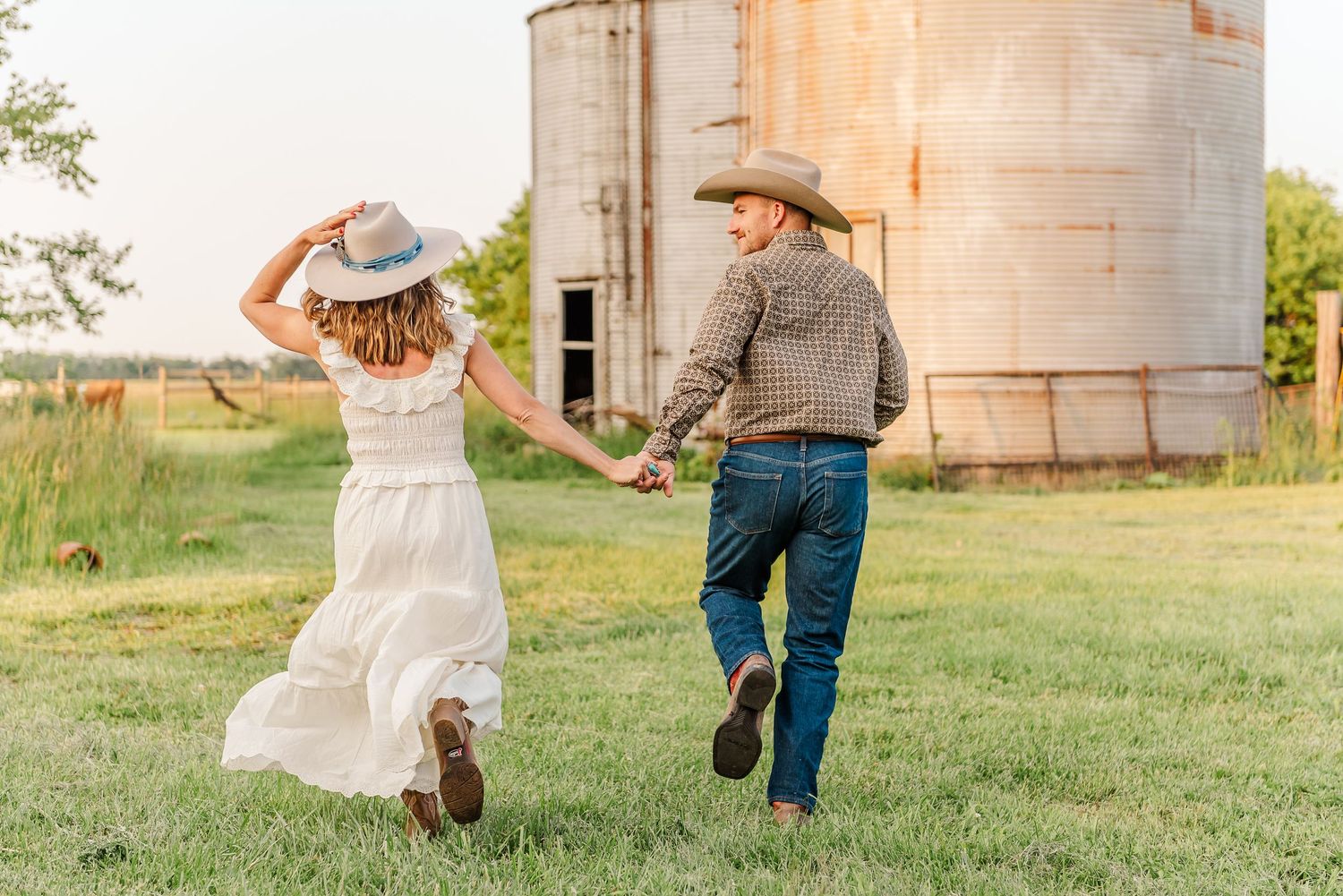 A couple walks hand in hand through a rustic farm setting with grain silos in the background during golden hour.