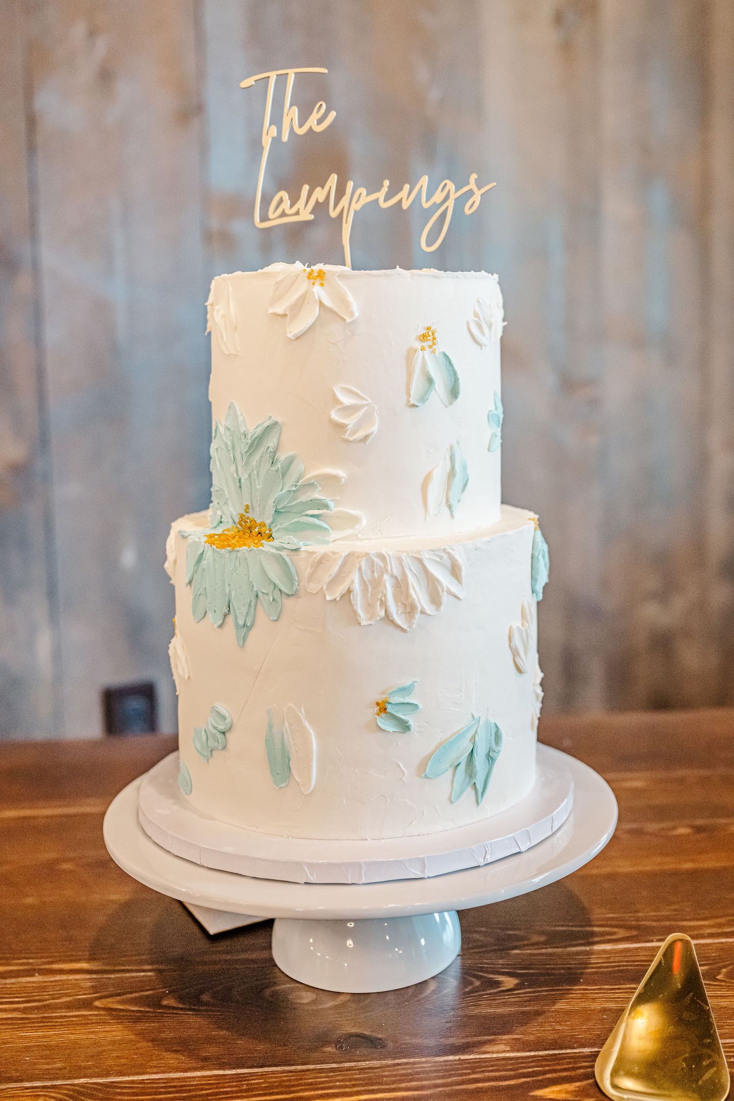 Elegant two-tier white wedding cake with blue floral designs and a gold 'The Lampings