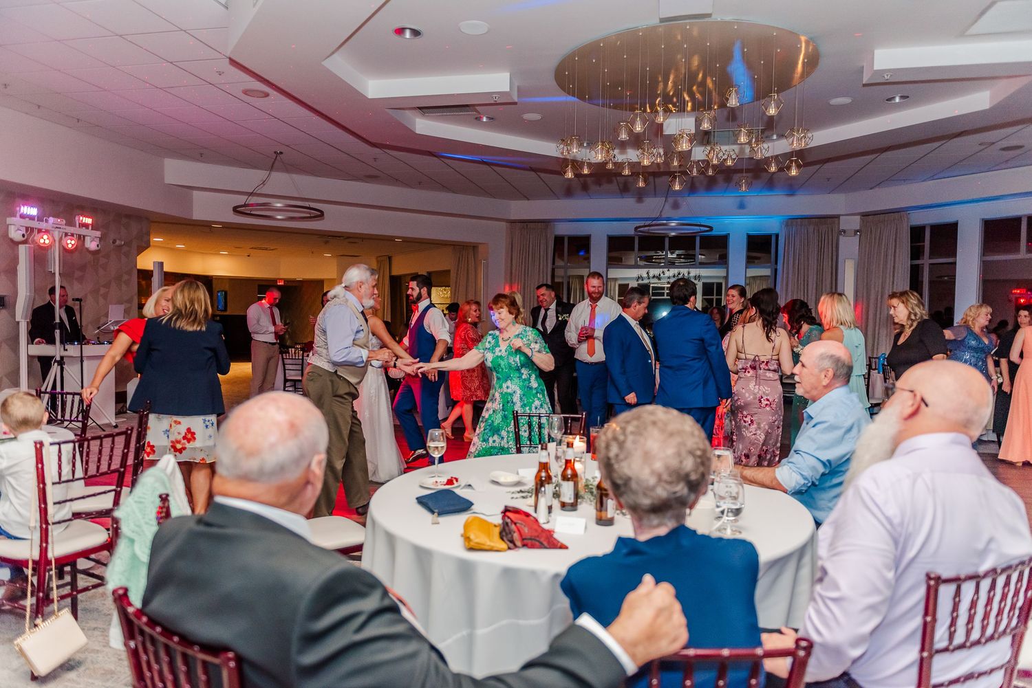 Wedding reception guests gather around tables in an elegantly decorated ballroom with pink and blue lighting.