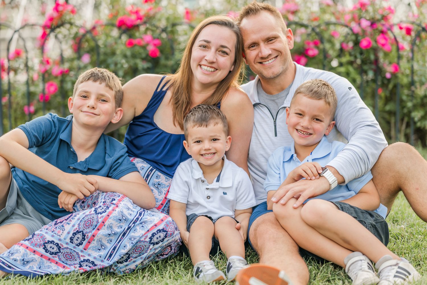 Happy family of five smiling outdoors on grass with pink flowers in background on a sunny day. Columbus Park of Roses.