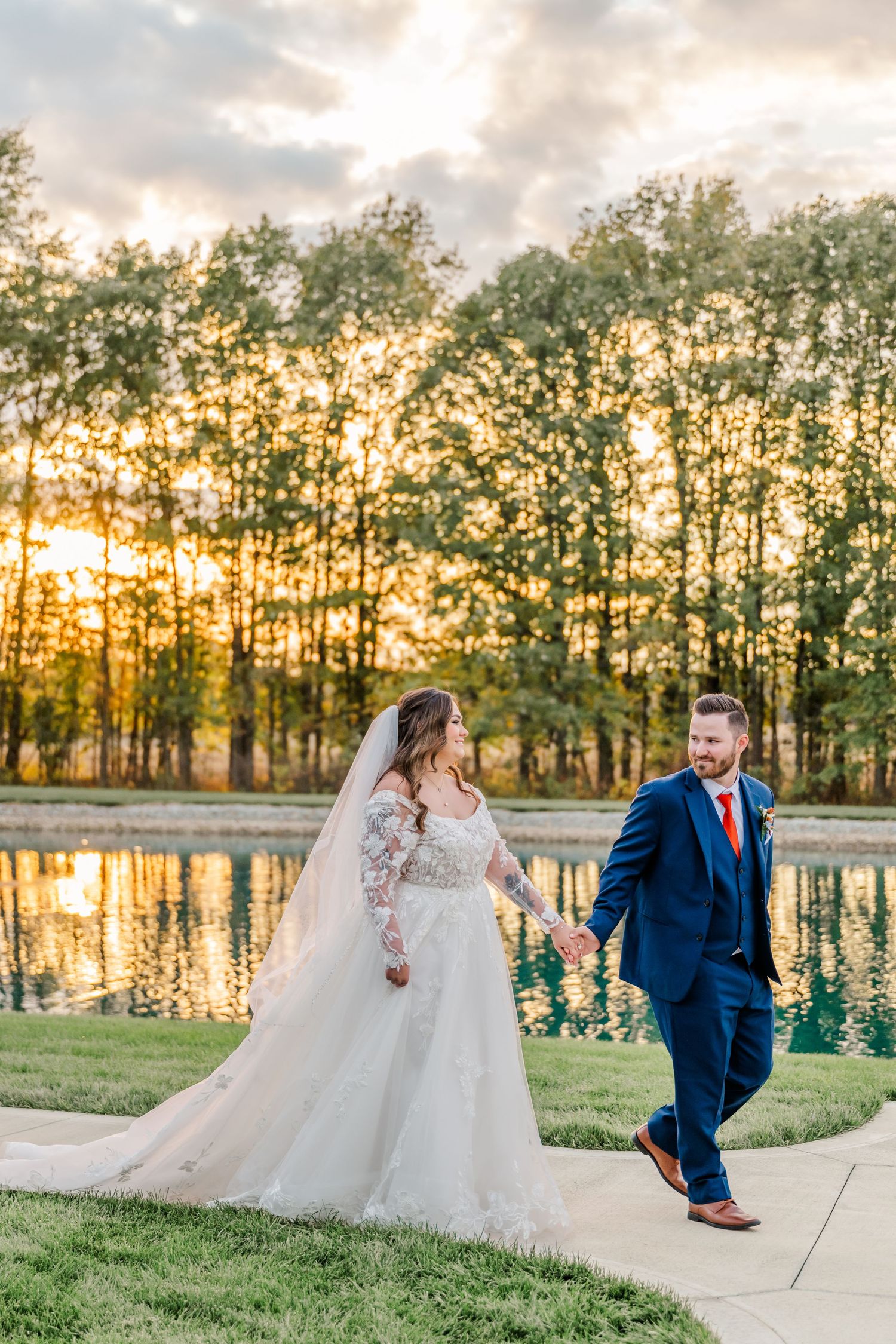 Couple poses at sunset by a serene lake with tall pine trees silhouetted against the golden sky.Four Seasons Barn Wedding & Event Venue