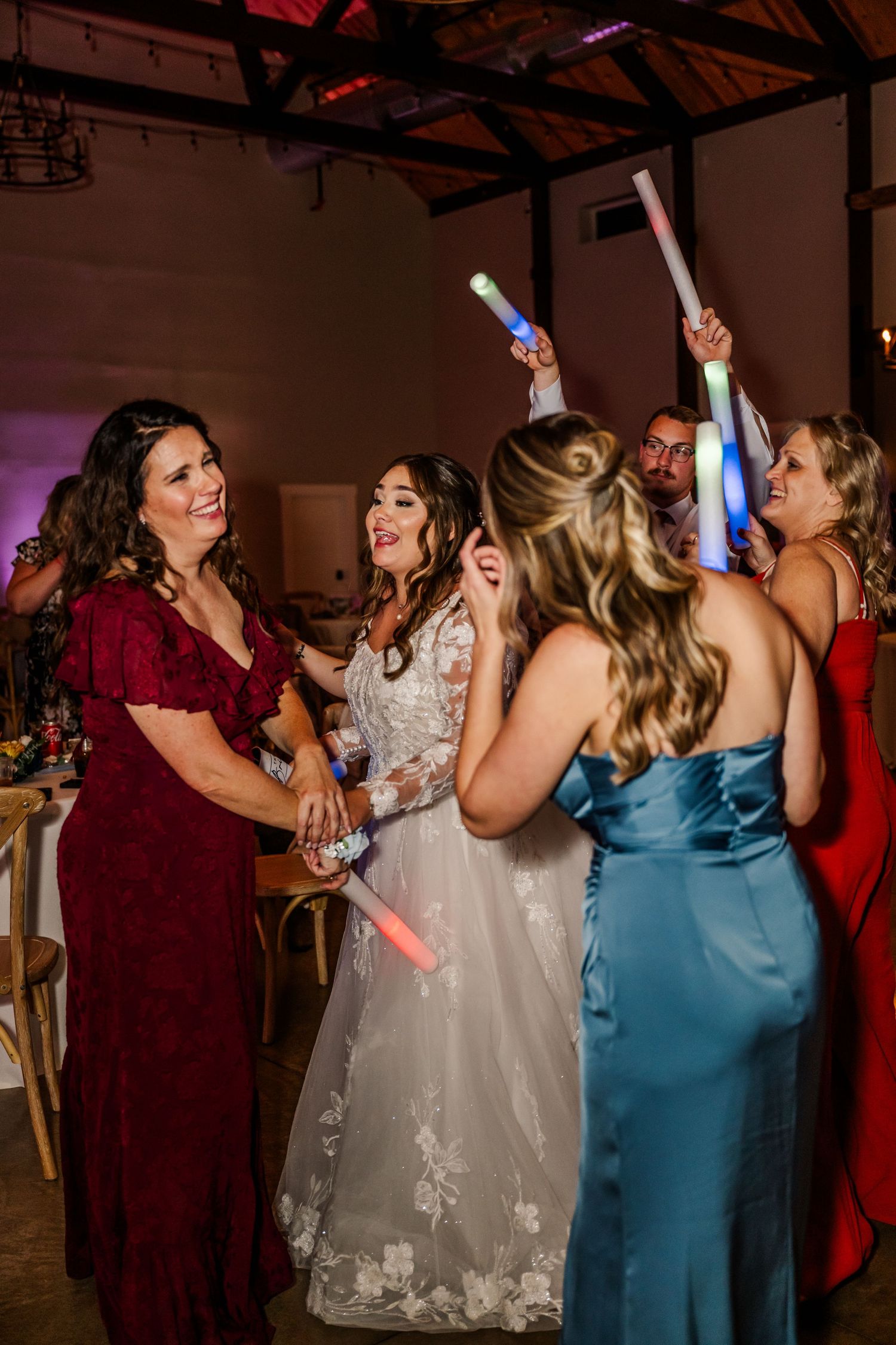 Wedding party celebrates on dance floor with light-up foam sticks at indoor reception venue.Four Seasons Barn Wedding & Event Venue