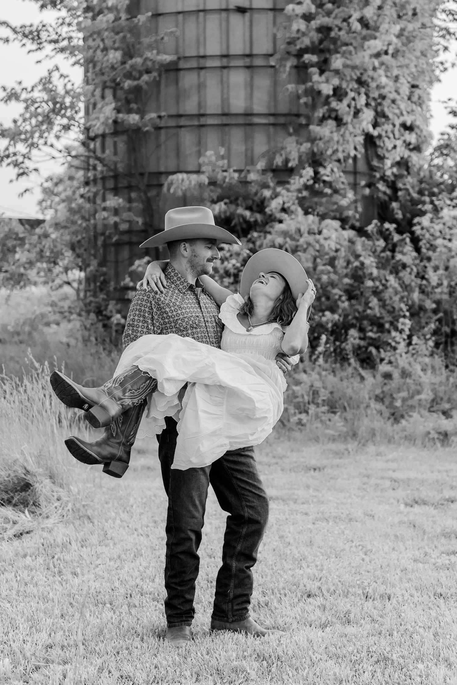 A romantic black and white photo of a couple in western attire playfully embracing near a rustic silo.