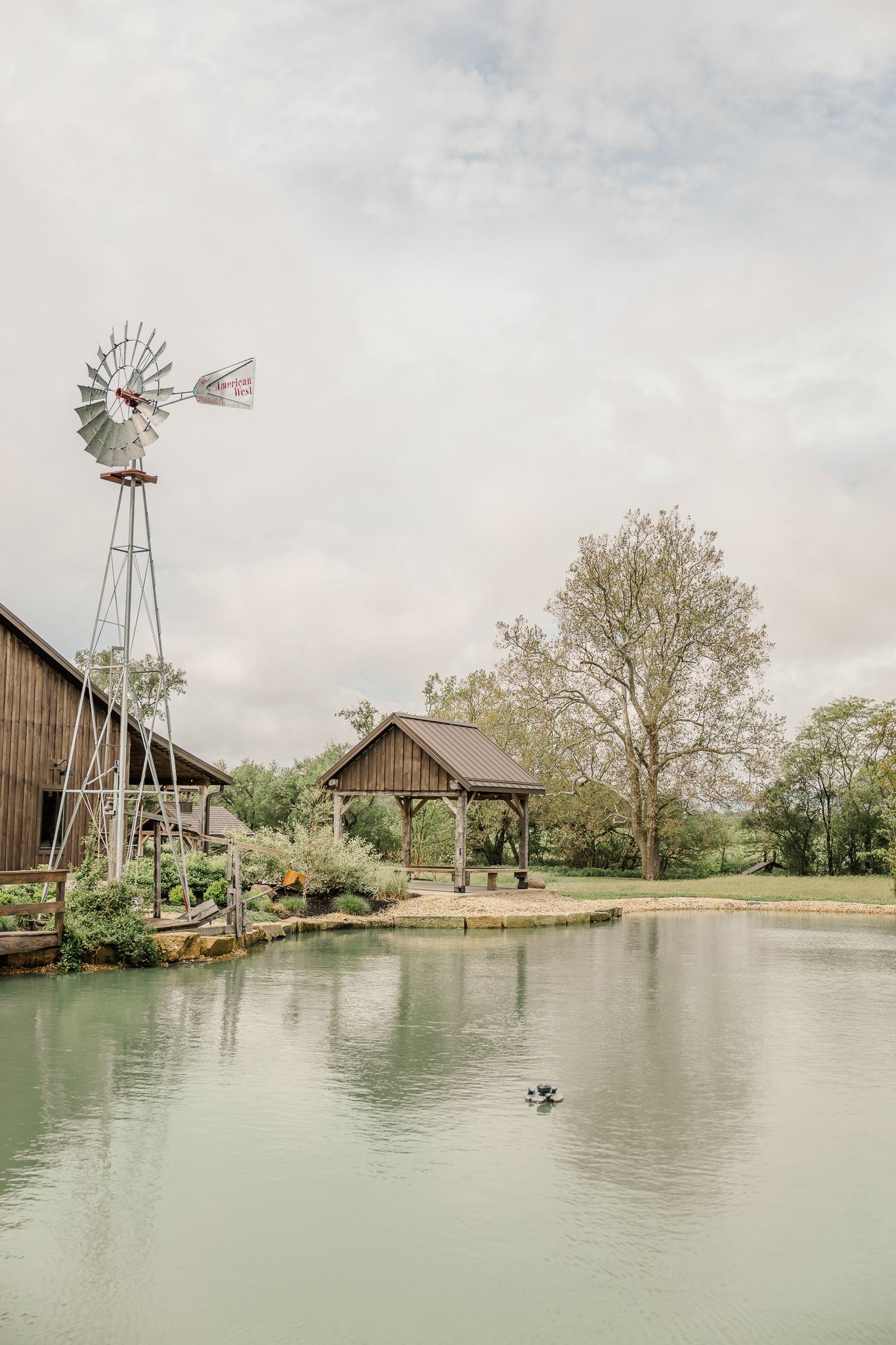 Rustic farm pond with a windmill, wooden gazebo, and lush trees reflected in calm water under a cloudy sky.The Old Barns at Dry Run Farms.