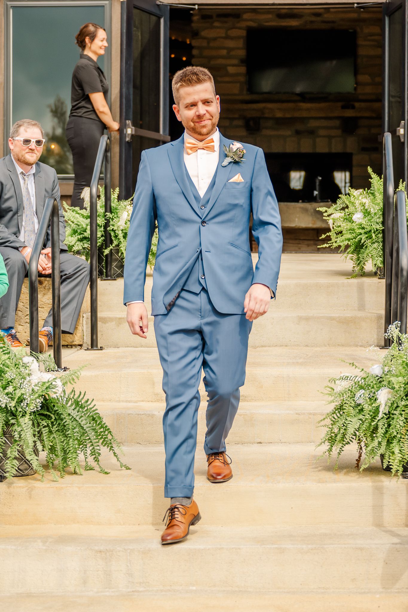 A groom in a blue three-piece suit with an orange bow tie walks down an outdoor aisle lined with fernsThe Old Barns at Dry Run Farms.