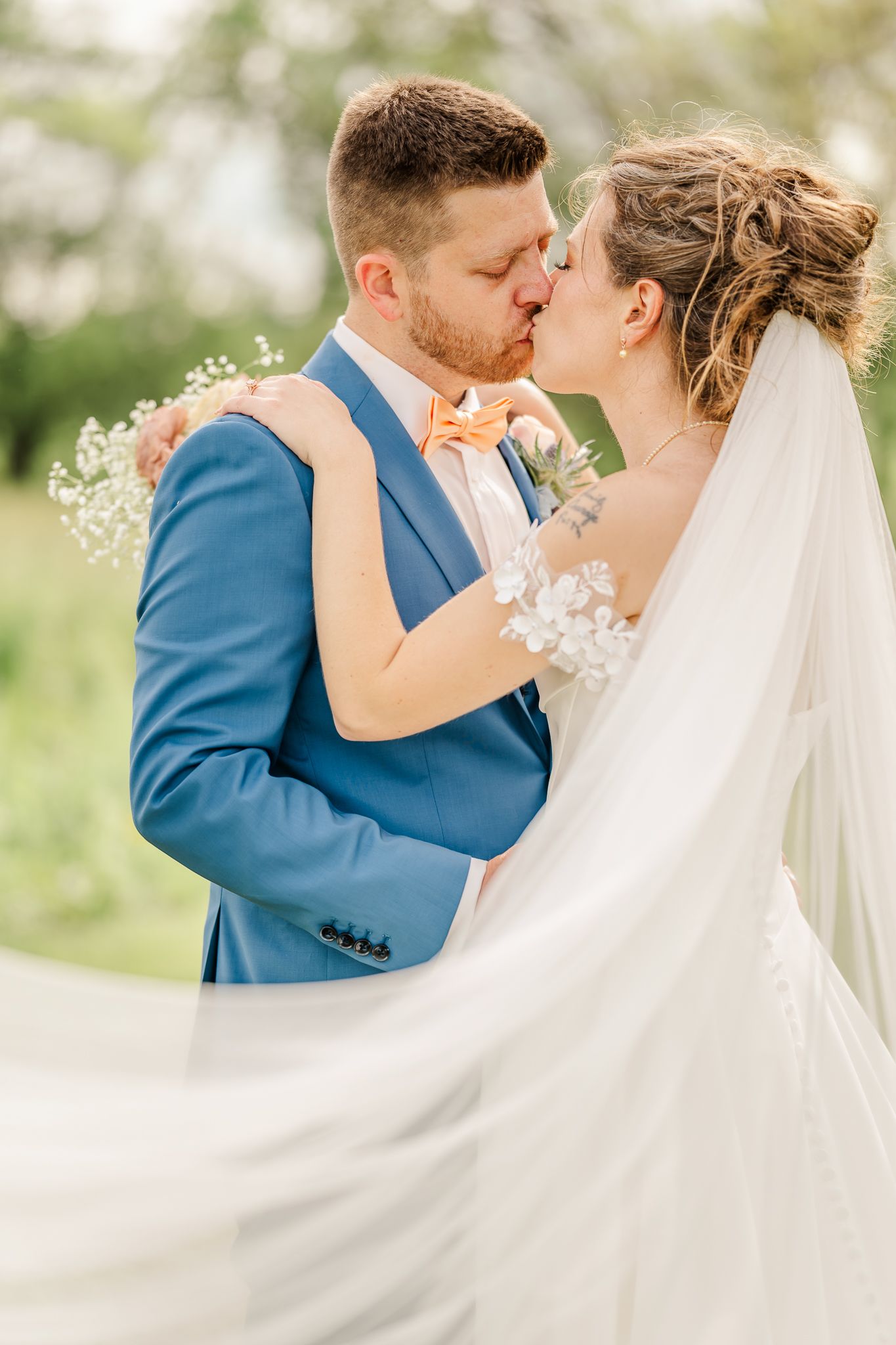 A bride and groom share a romantic kiss outdoors, the groom in a blue suit and the bride in a white gown with a flowing veil.