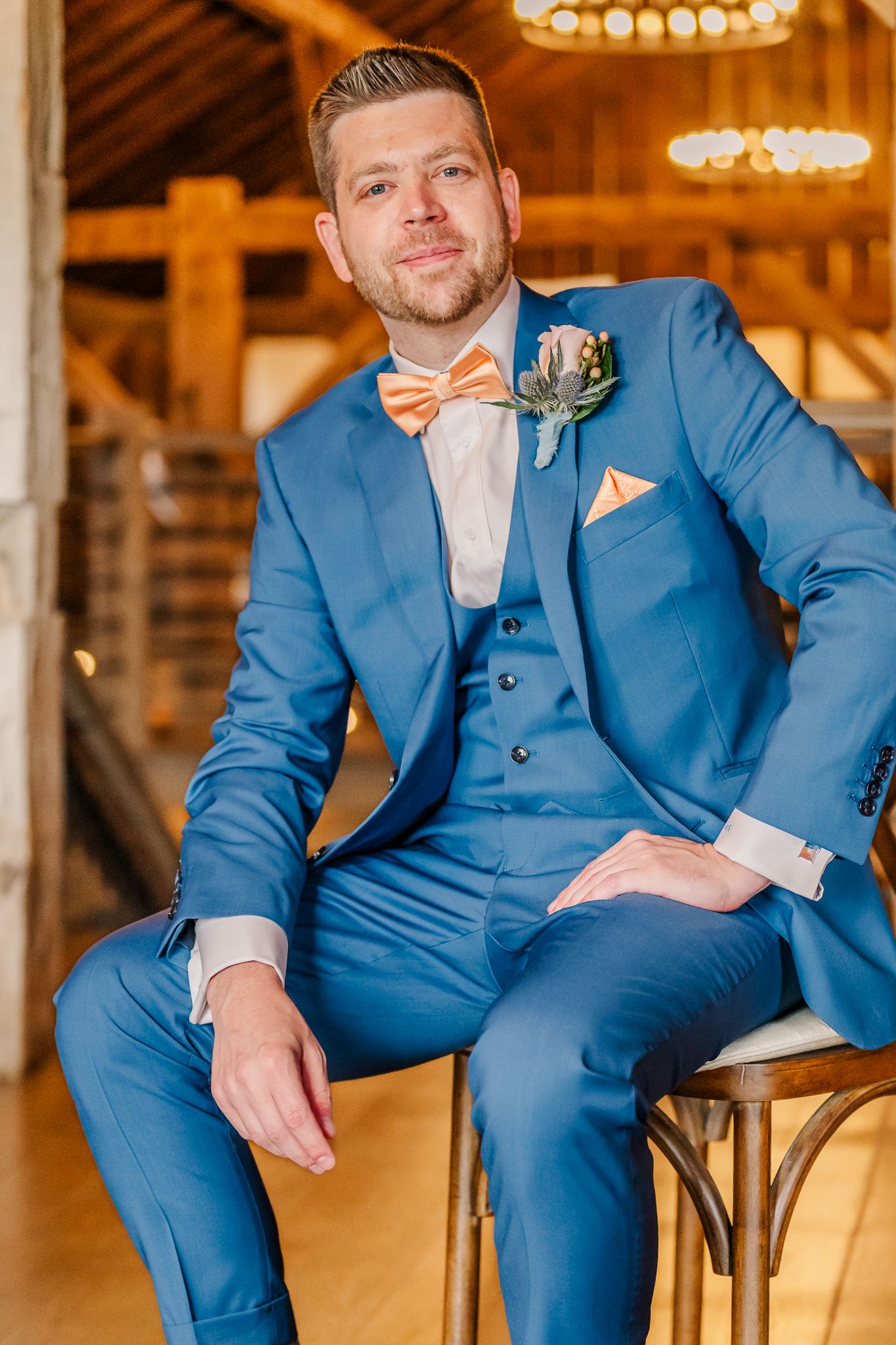 Groom in blue three-piece suit with orange bow tie and boutonniere sits confidently in a rustic barn venueThe Old Barns at Dry Run Farms.