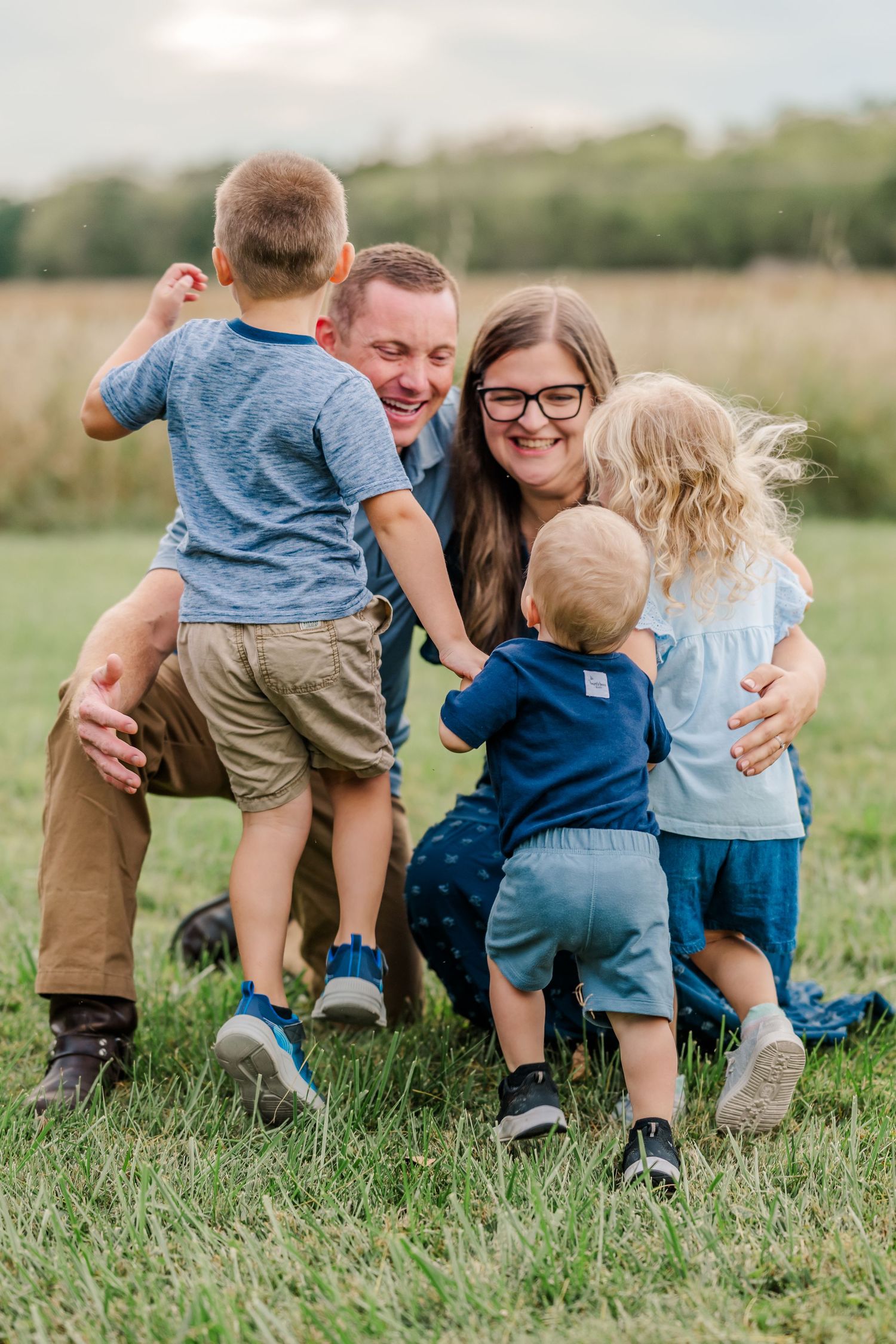 A joyful family of five plays together on a green field, with parents laughing as three young children gather around them.