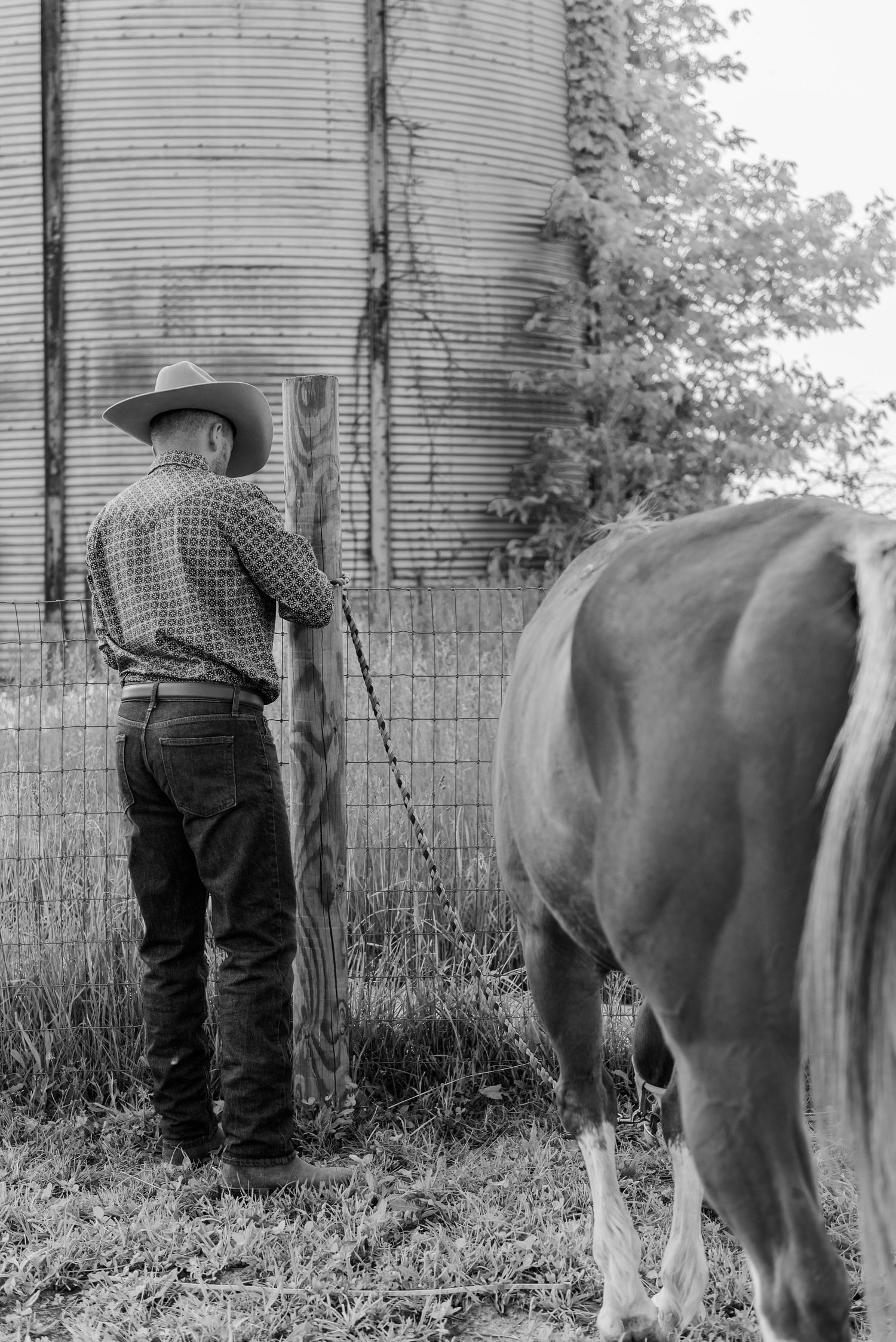 Black and white series showing a person in western wear standing with a horse near a grain silo.