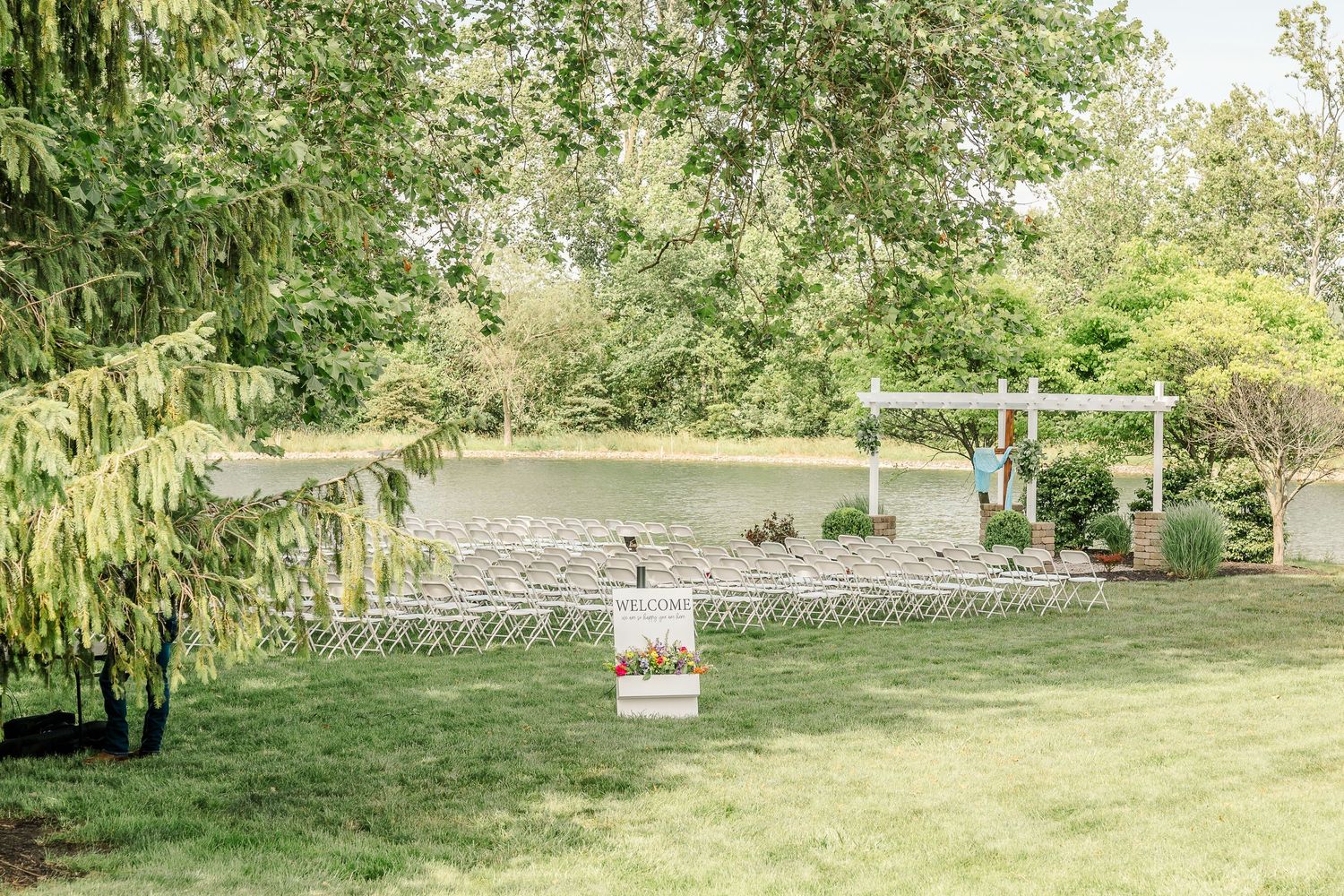 Outdoor wedding ceremony setup with white chairs and an arbor overlooking a serene pond surrounded by willow trees.
