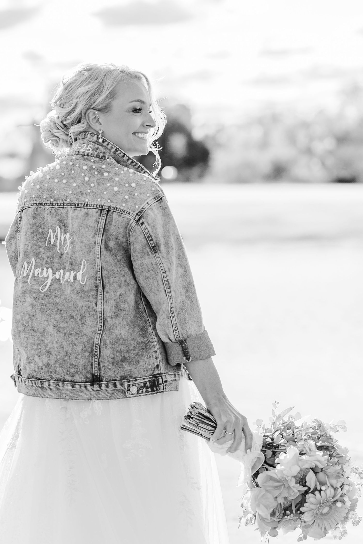 A bride in a customized denim jacket over her wedding dress holds a bouquet while looking over her shoulder.