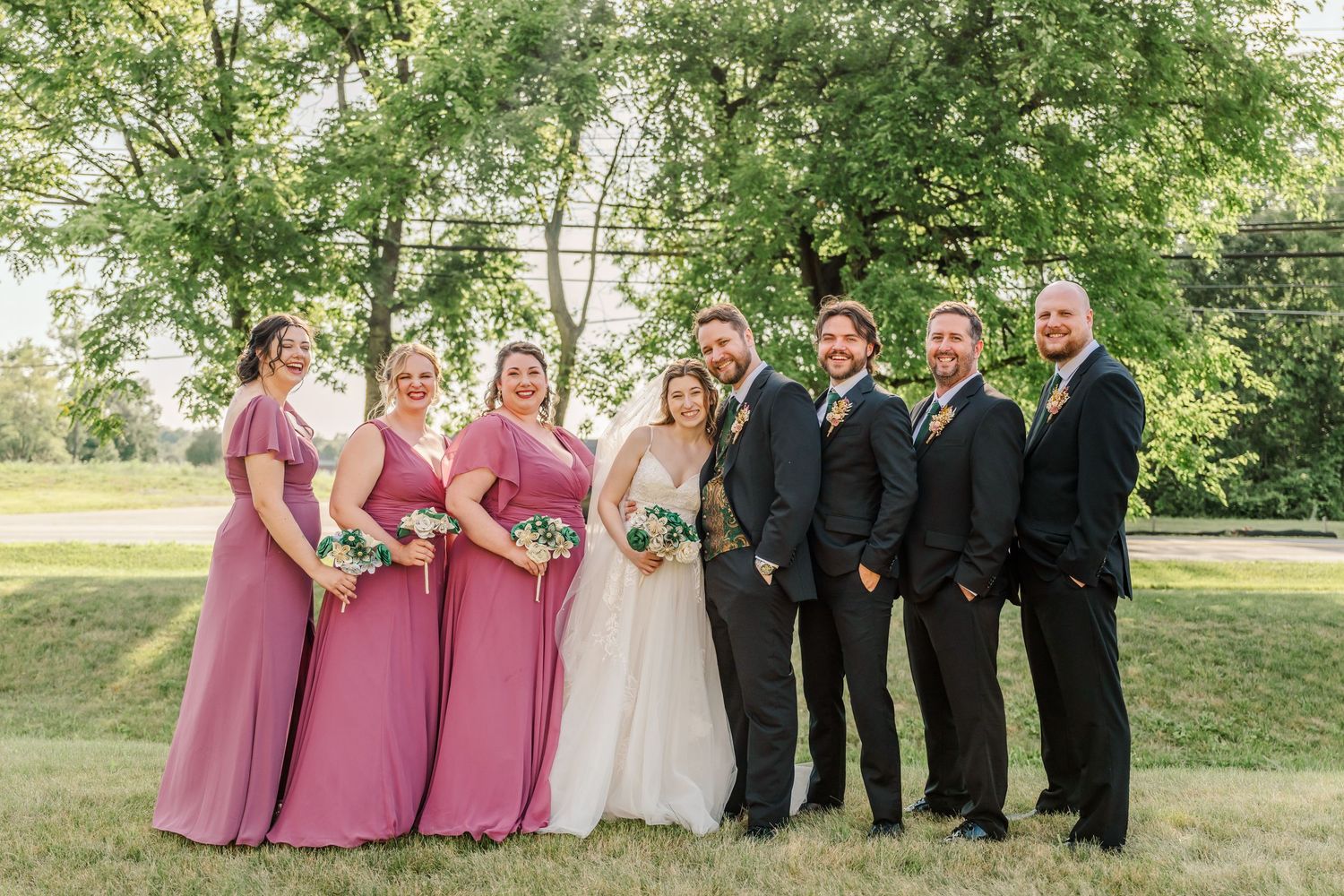 Wedding party in pink dresses and black suits posing outdoors in a garden setting.