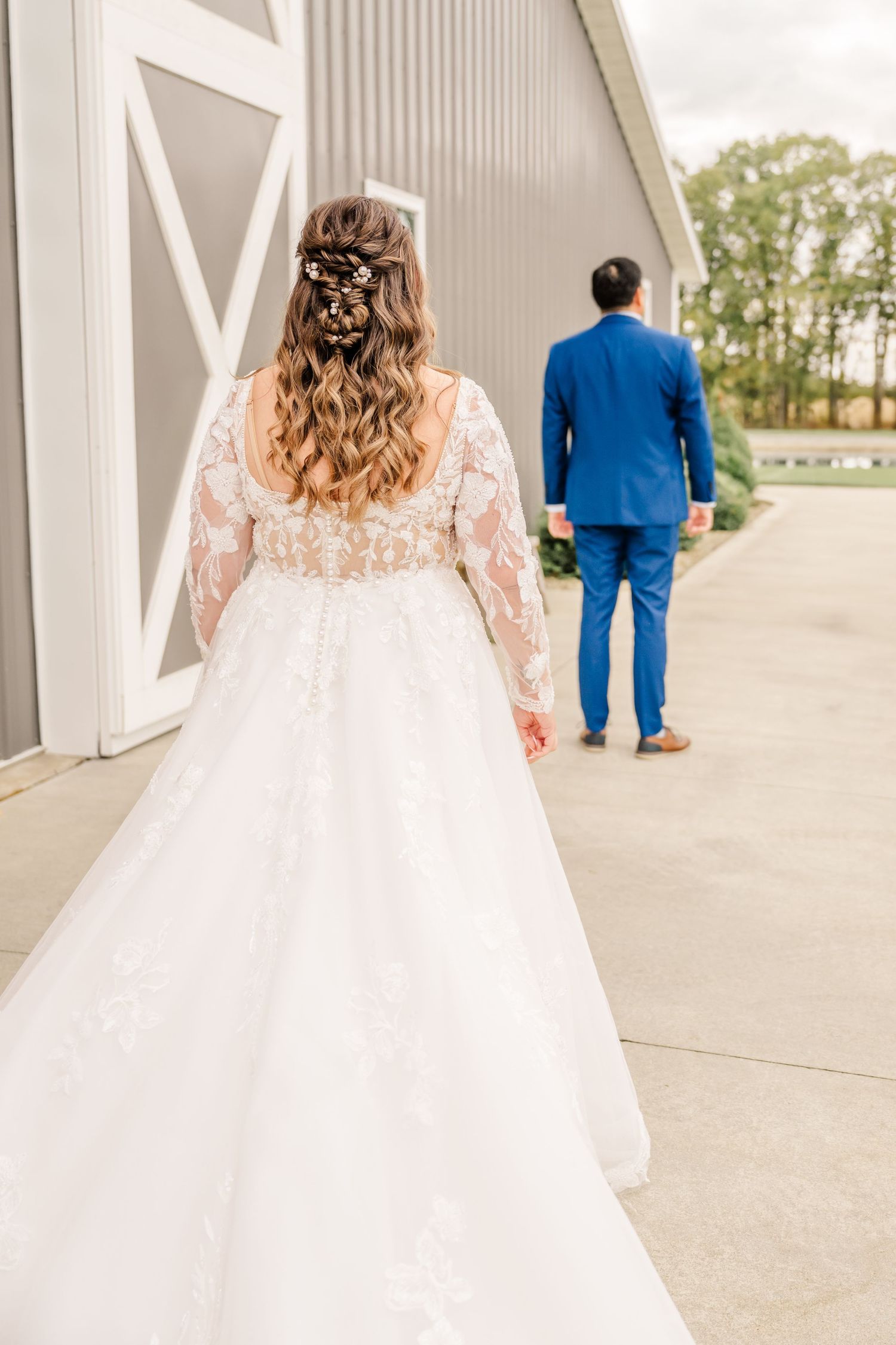 A bride in a lace wedding dress walks behind a groom in a blue suit along a covered walkway.