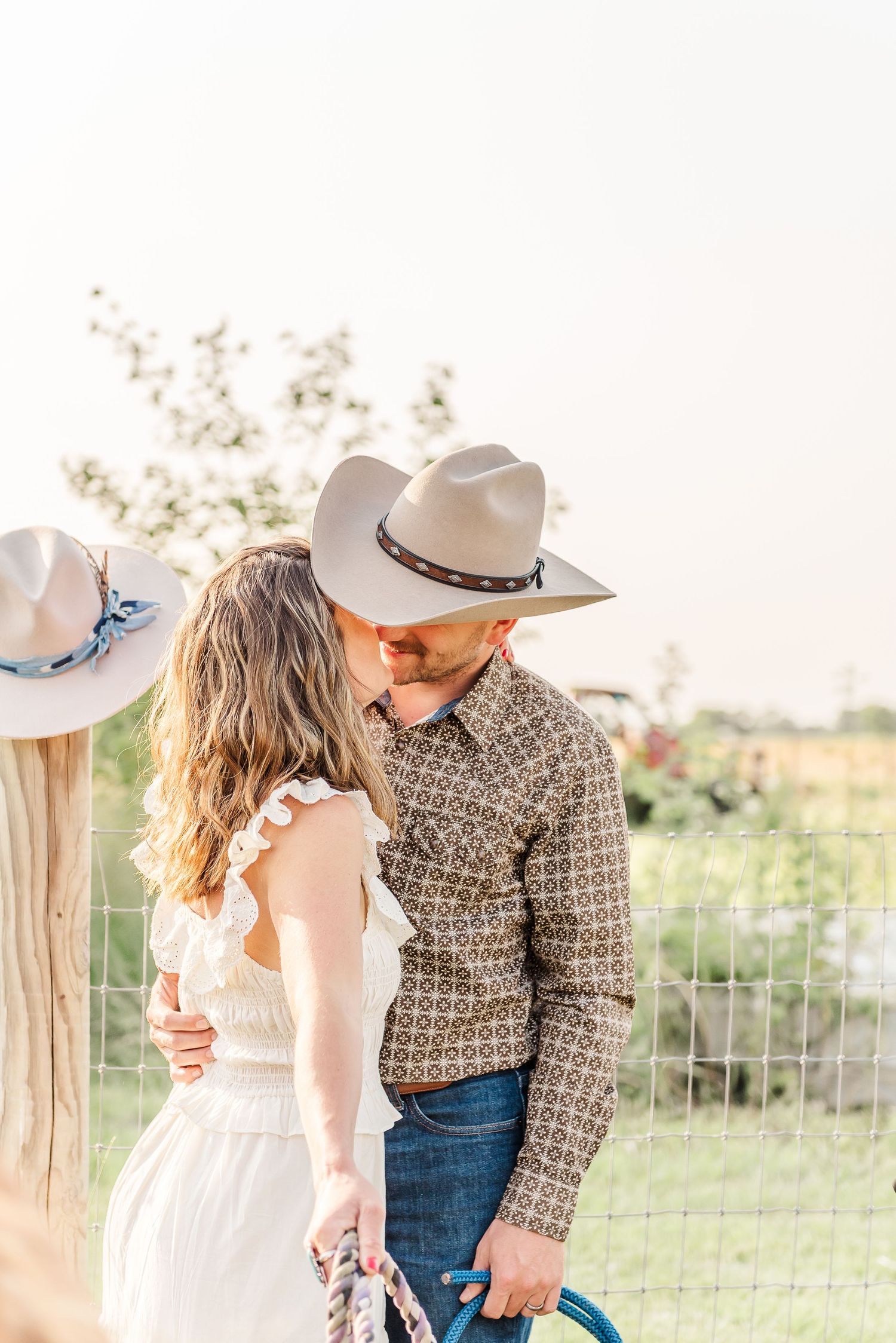 A romantic couple in western attire embraces in a sunlit field during a golden hour photoshoot.