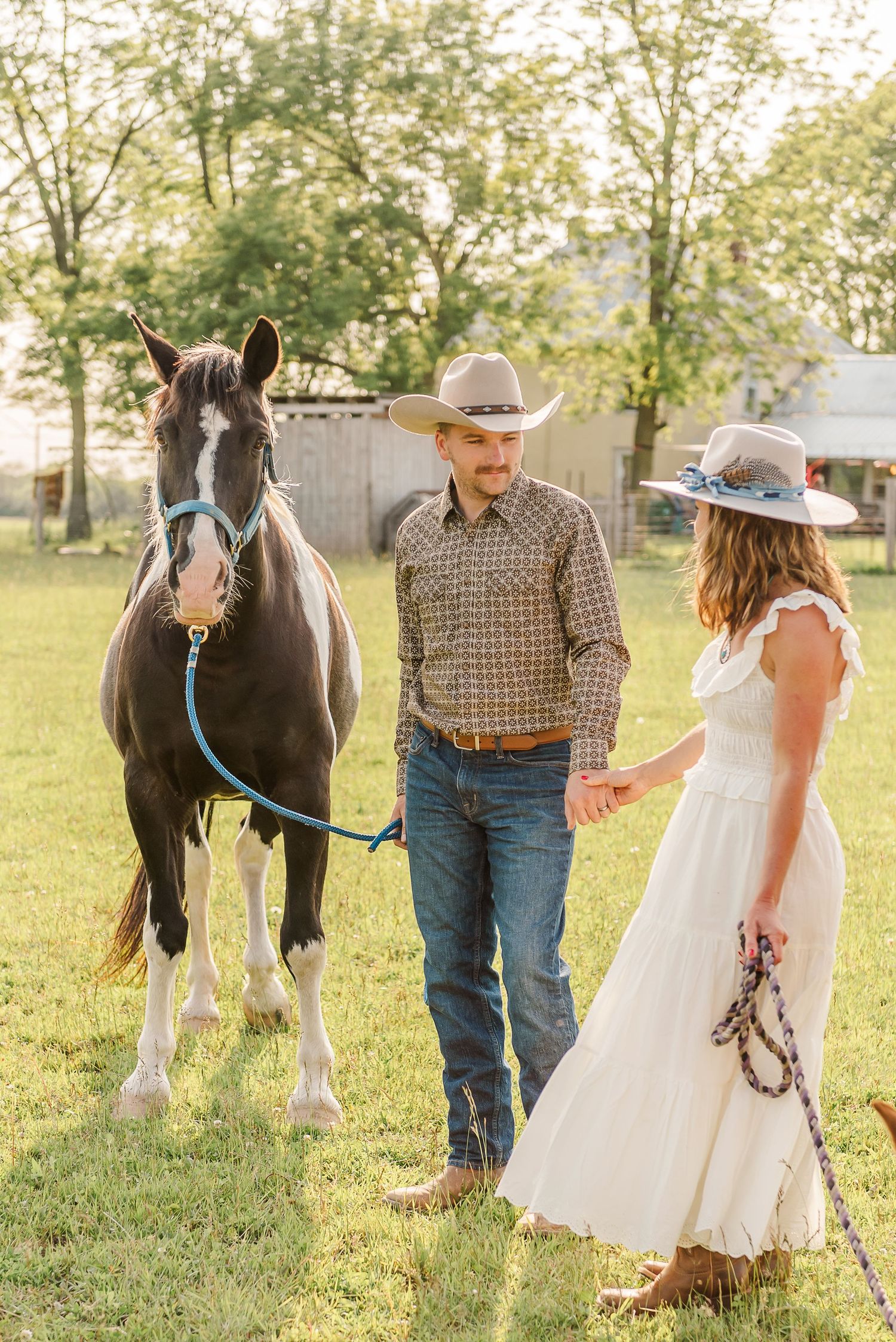 A couple in western attire poses with a black horse in a sunlit field with trees in the background.