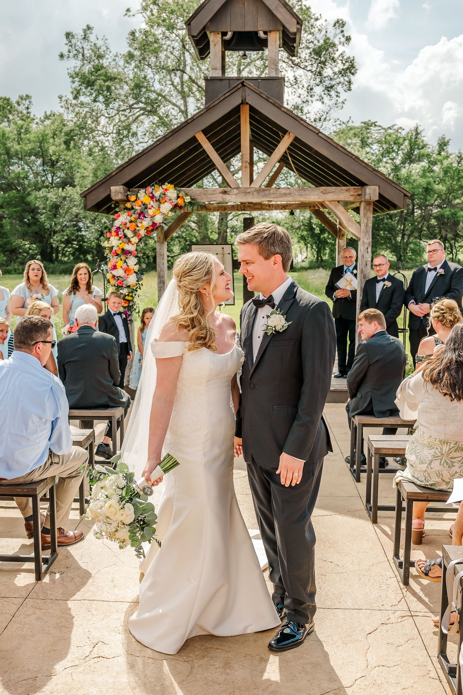 Guests gather at an outdoor wedding ceremony under a wooden pavilion with colorful floral decorations.The Old Barns at Dry Run Farms