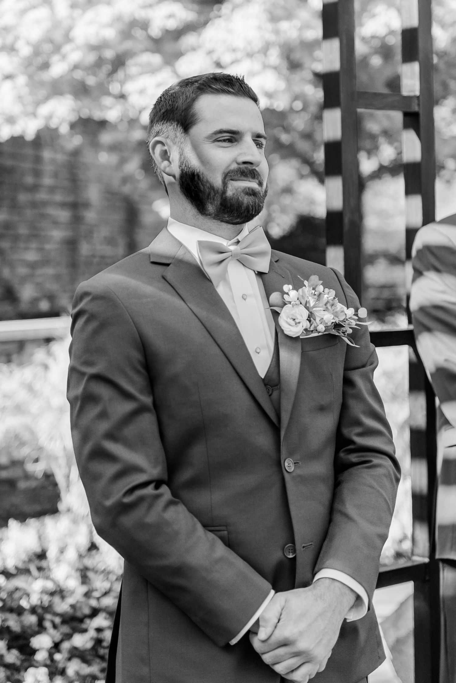 A groom in a dark suit and tie stands at the altar during an outdoor wedding ceremony.