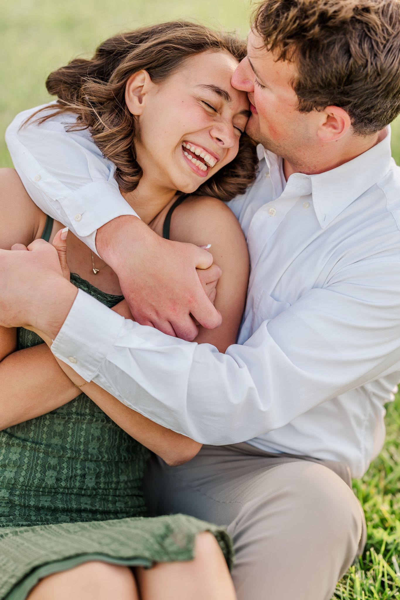 A joyful couple sitting on grass, man in white shirt kissing woman's cheek as she laughs happily. The Old Barns at Dry Run Farms.
