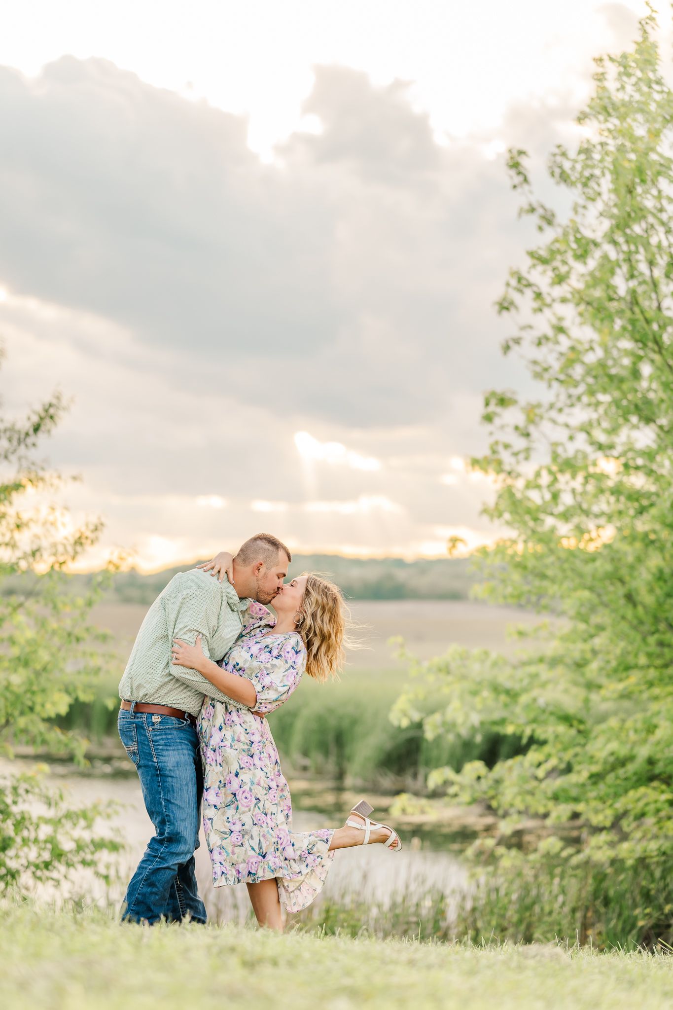 Couple sharing a romantic kiss outdoors in a lush green field with a pond and dramatic cloudy sky at golden hour.