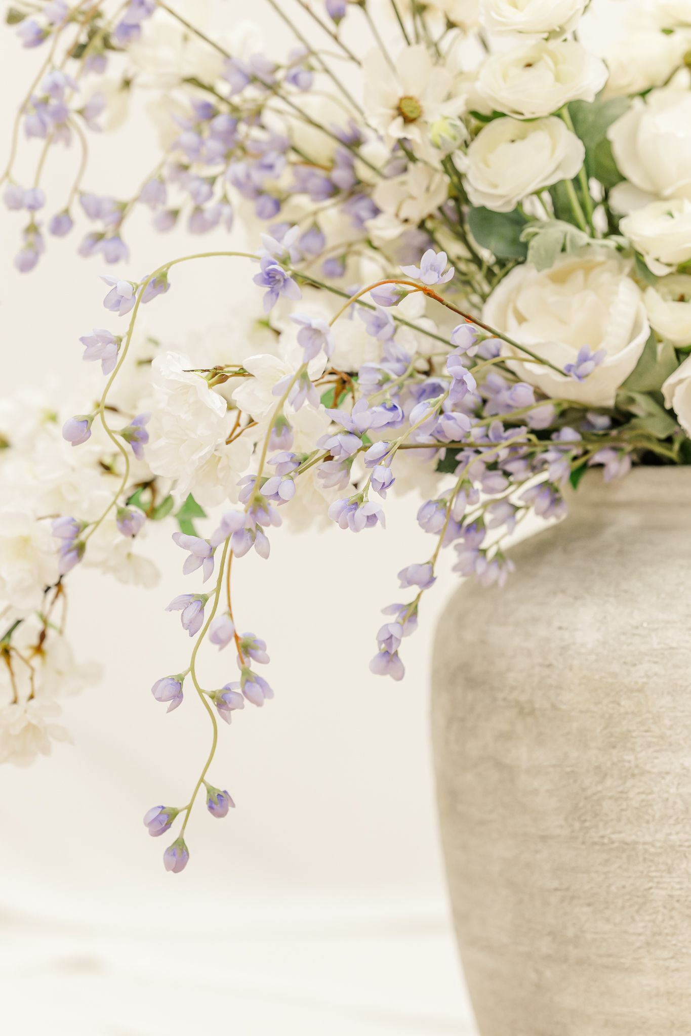 Delicate white and purple wisteria flowers arranged in a light gray ceramic vase against a soft neutral background.