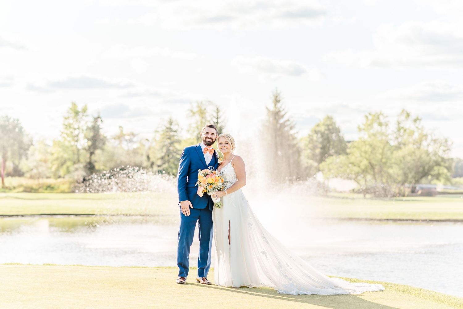 A couple in wedding attire stands together on a sunlit golf course with a lake and trees in the background.