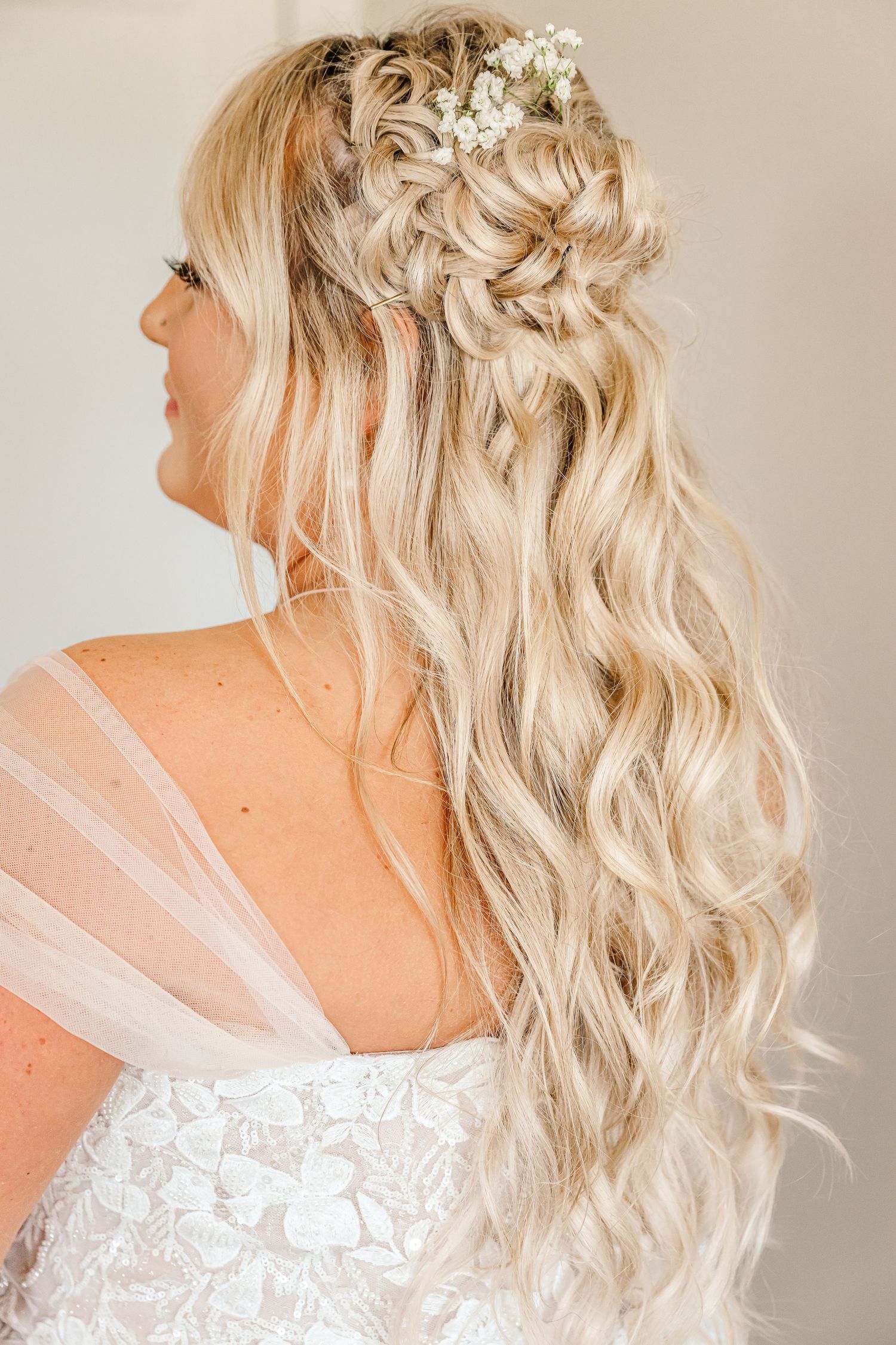 Elegant bridal hairstyle featuring cascading blonde waves with delicate white floral accessories against light background.