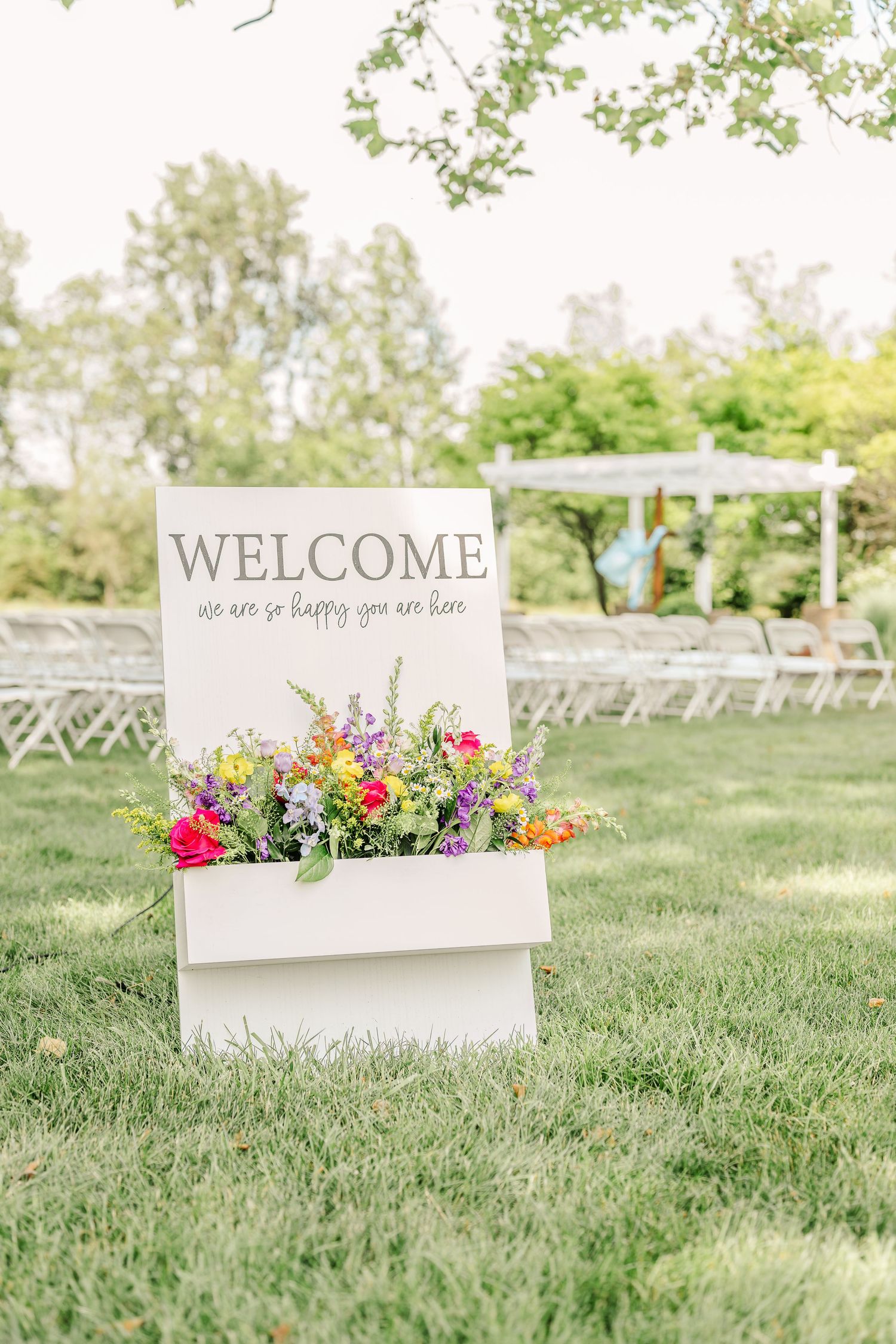 White welcome signs with colorful floral arrangements displayed on a grassy lawn at an outdoor wedding venue.