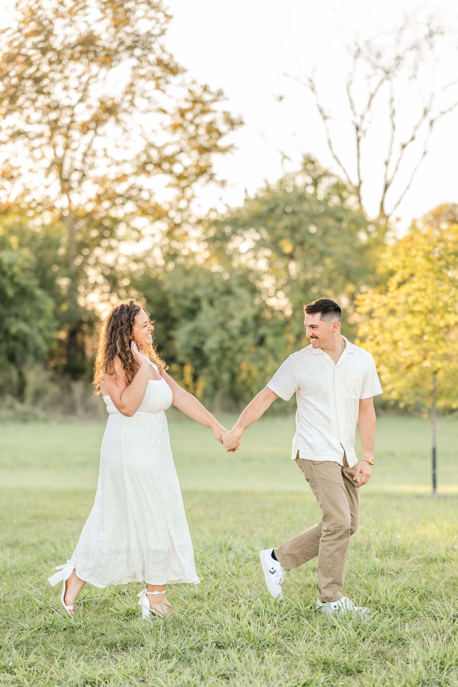 Smiling couple in white outfits holding hands and walking through a sunlit grassy field with trees in the background.