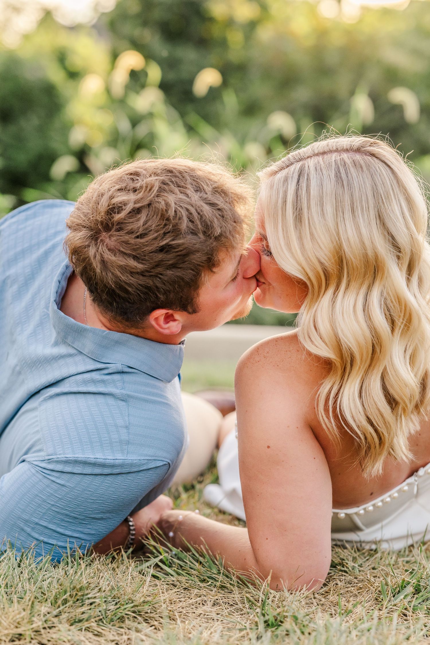 Romantic couple lying in grass wearing casual blue and white outfits share tender kisses in outdoor photoshoot sequence.