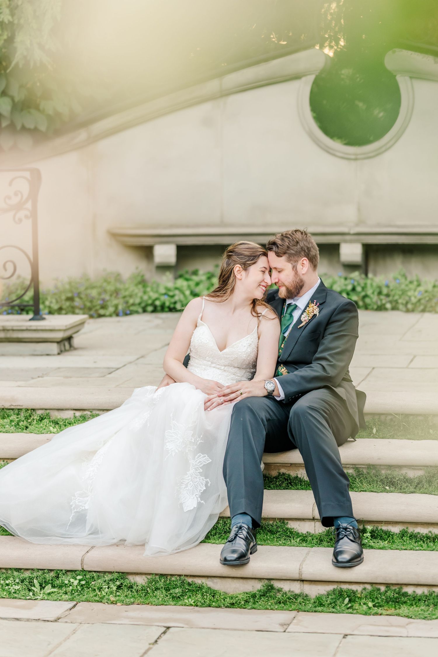 Romantic wedding couple resting on stone steps surrounded by garden landscaping.
