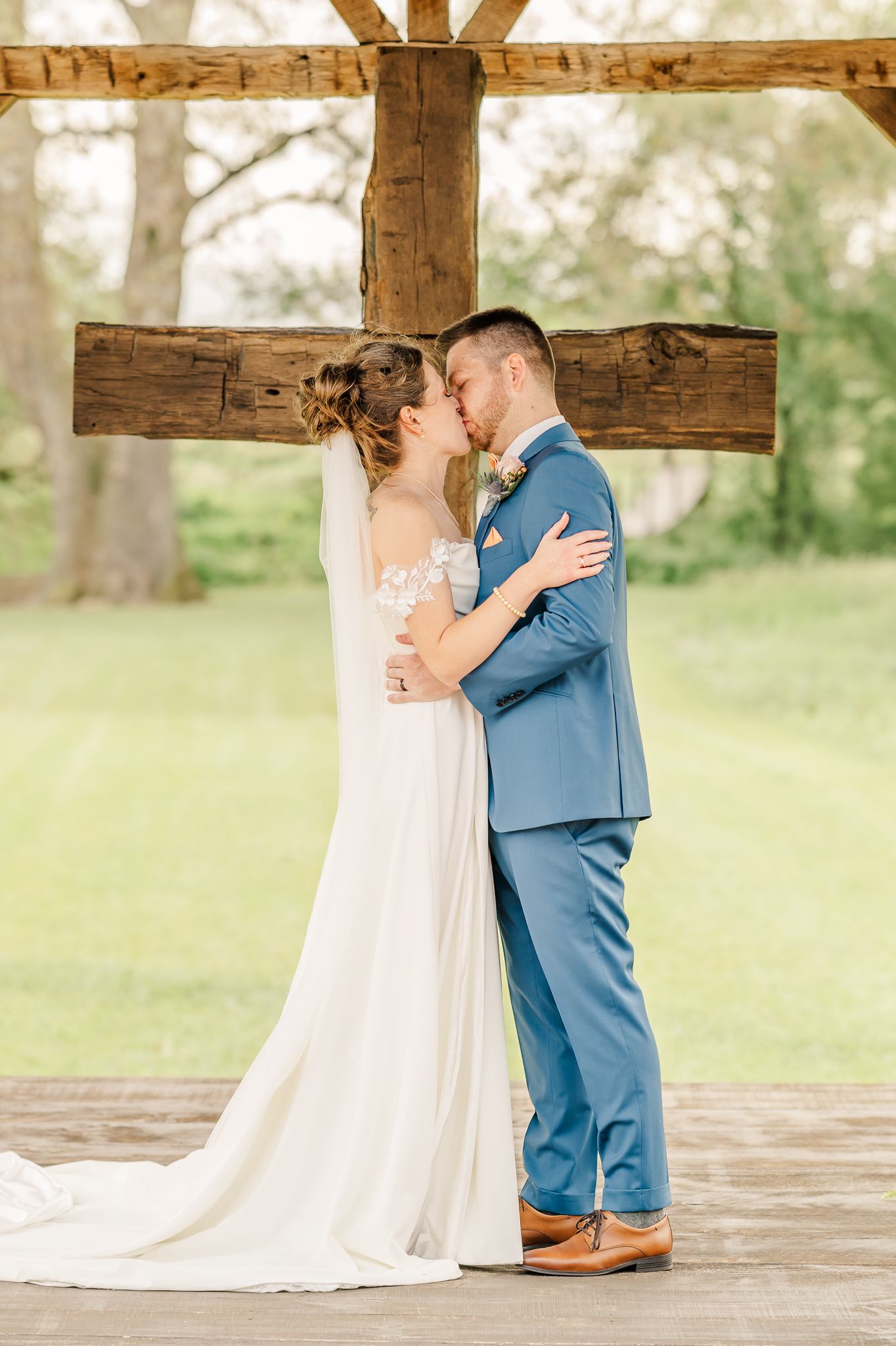 Bride and groom share a romantic kiss in front of a rustic wooden cross outdoors on their wedding day.The Old Barns at Dry Run Farms in Ohio