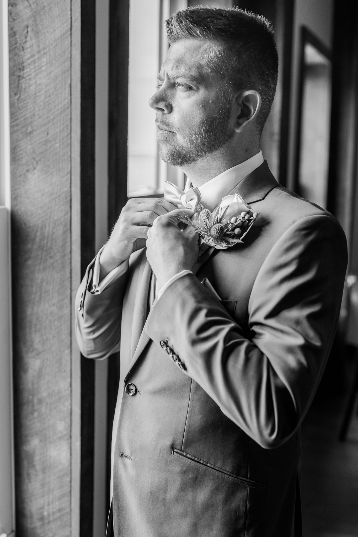 A groom in a suit adjusts his boutonniere and bow tie while standing by a window in a black and white photo.
