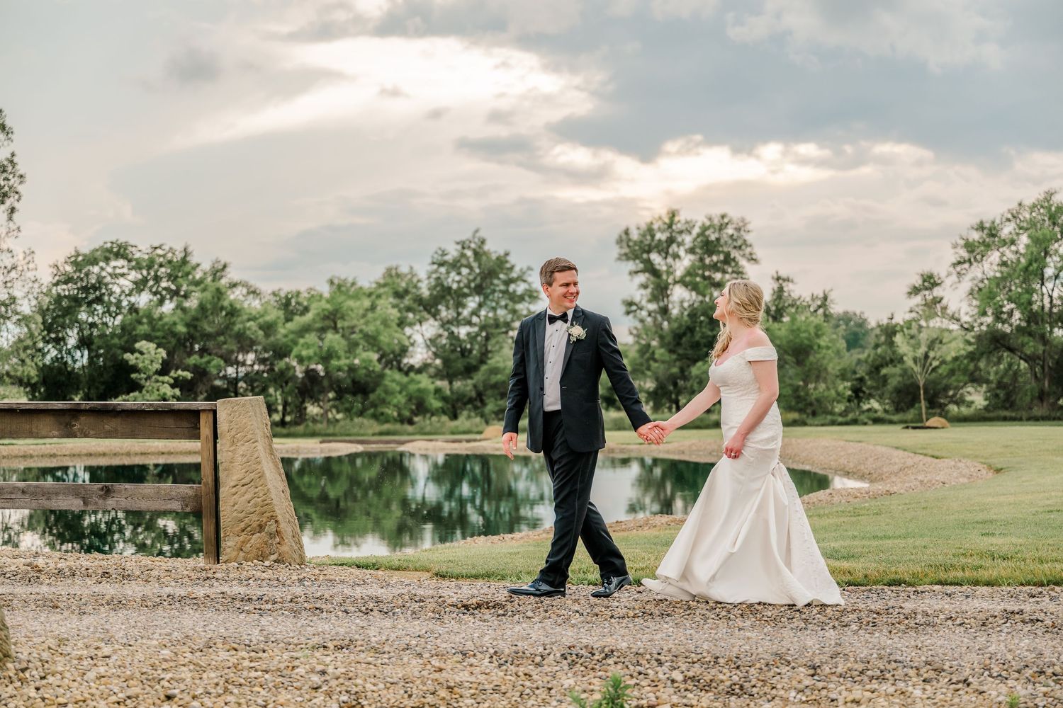 A couple walks hand in hand along a gravel path beside a serene pond on their wedding day.The Old Barns at Dry Run Farms in Williamsport, OH