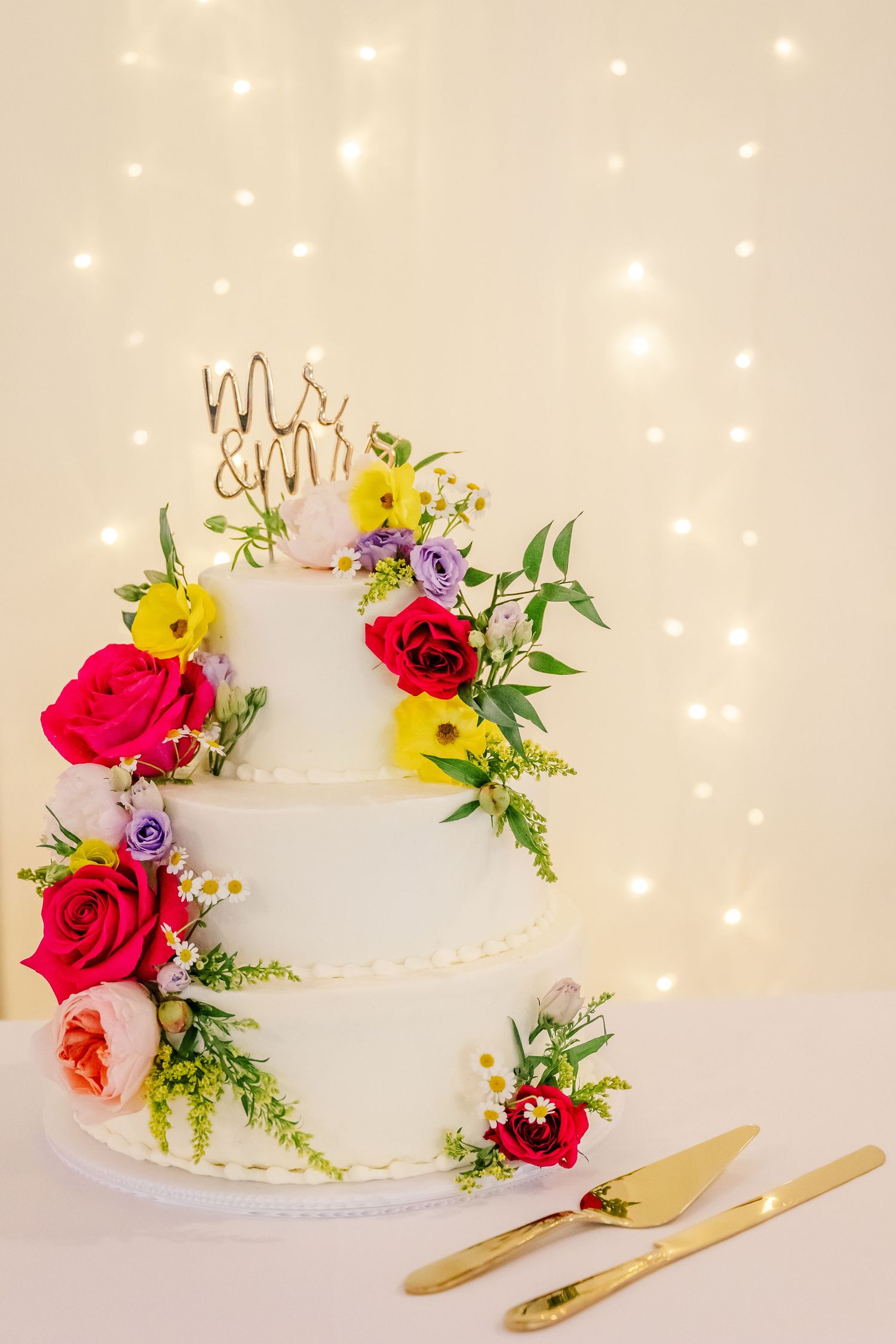 Three tiered white wedding cake decorated with vibrant red and yellow roses on a white surface with twinkling lights background.