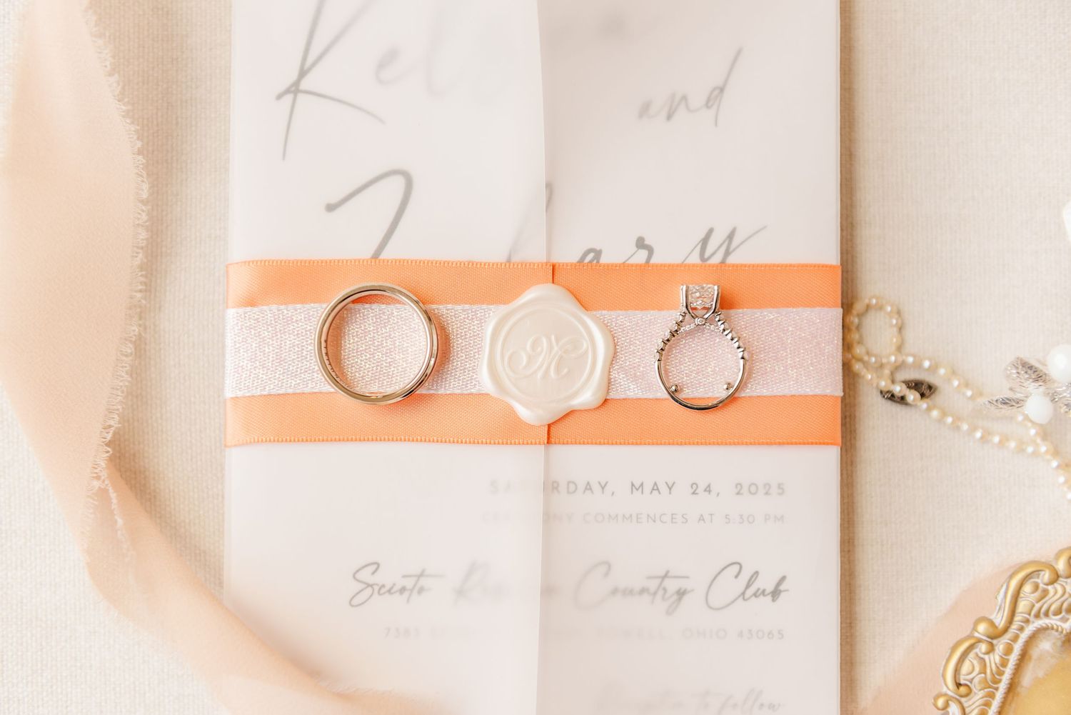 Wedding invitation suite with peach accents, rings, and delicate paper details laid out on white surface.