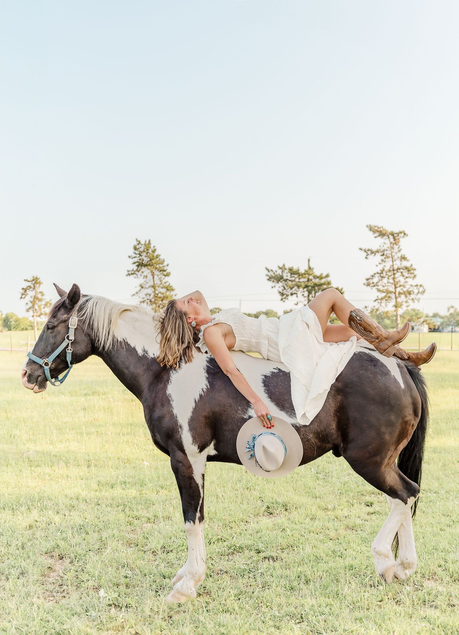 A person in a cowboy hat lies draped backwards across a black and white paint horse in a sunny field.