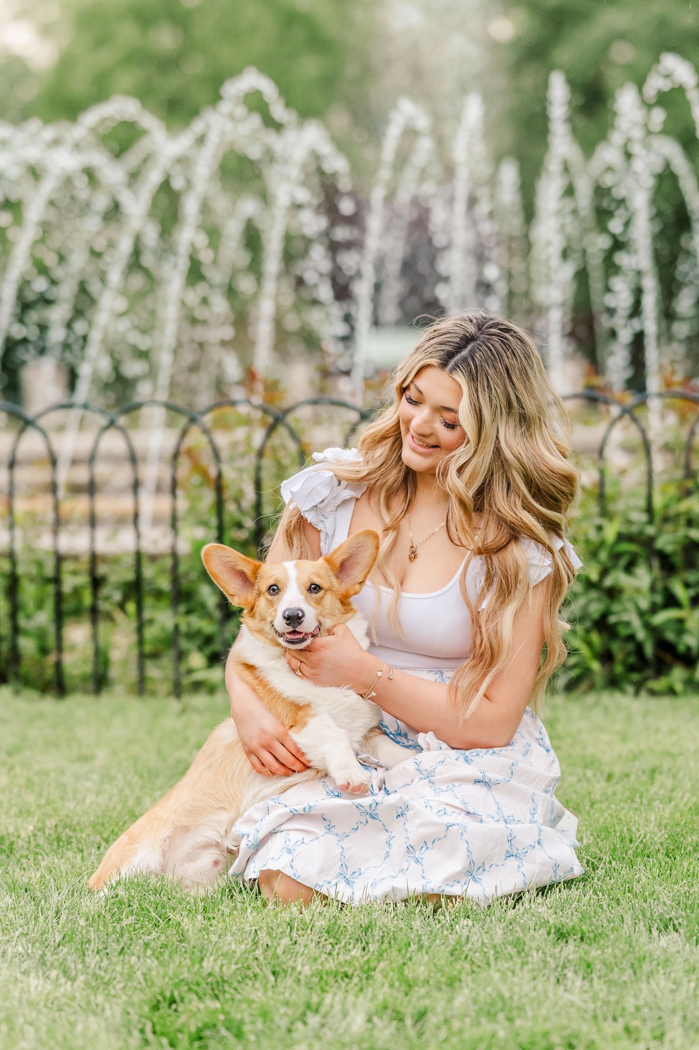 Young woman in floral skirt sitting on grass with a Corgi dog near decorative fountain in park. Columbus Park of Roses.