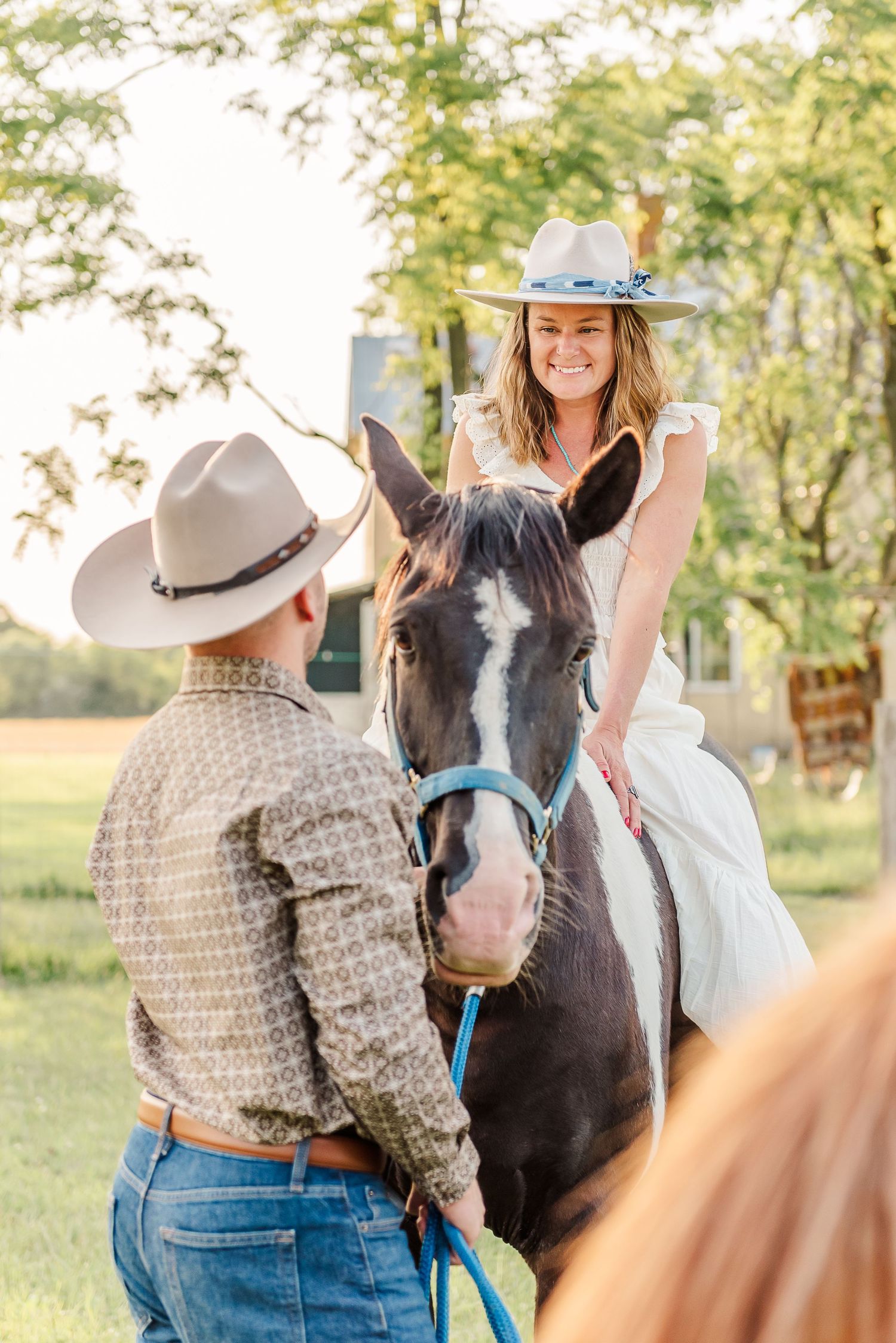 A couple in western attire poses with their paint horse in a sunny field during a photo session.