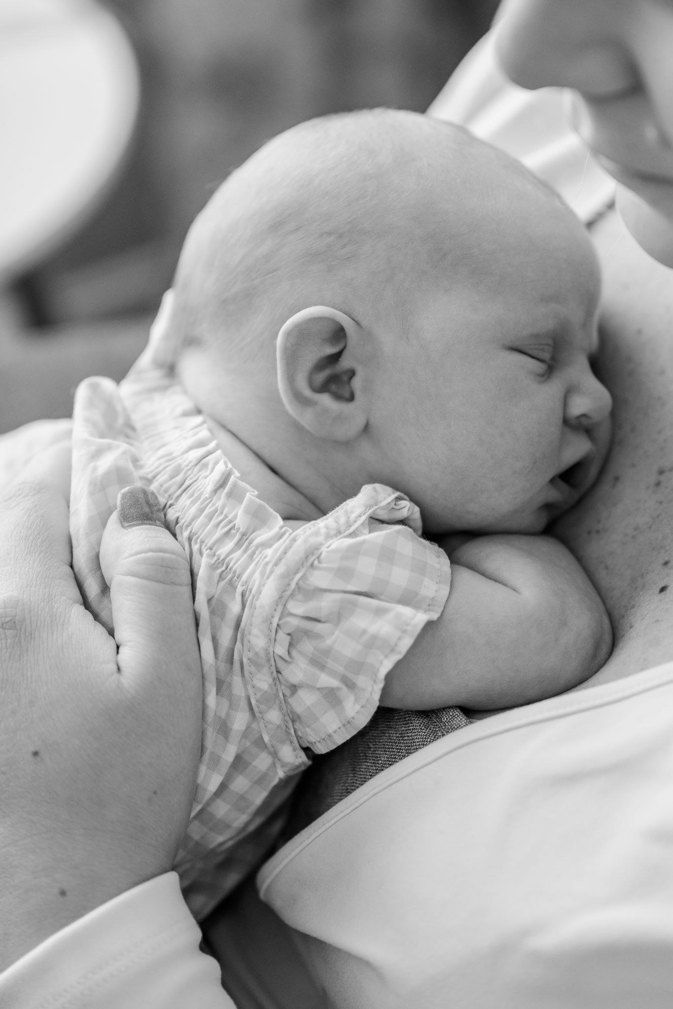 Black and white photo of a sleeping newborn baby cradled gently in adult hands, wearing a gingham outfit.
