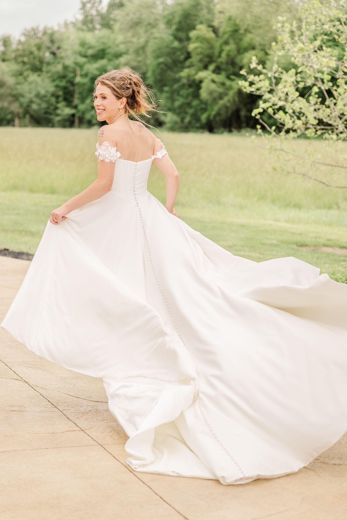 Bride twirling in elegant off-shoulder white ball gown with floral details outdoors on a sunny day. The Old Barns at Dry Run Farms.