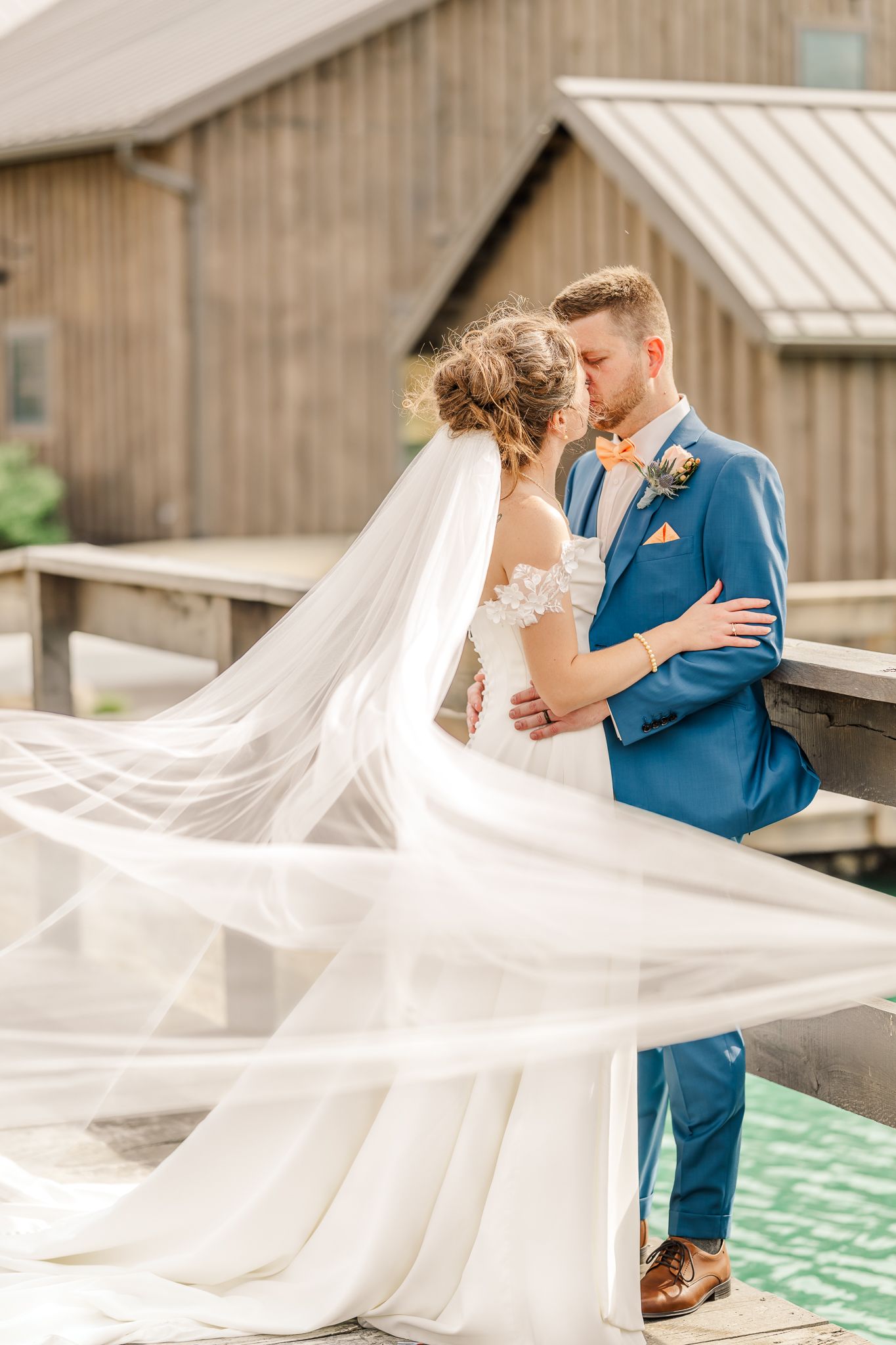 Bride and groom share a romantic kiss on a wooden dock, her veil flowing in the wind, groom in a blue suit. The Old Barns at Dry Run Farms.