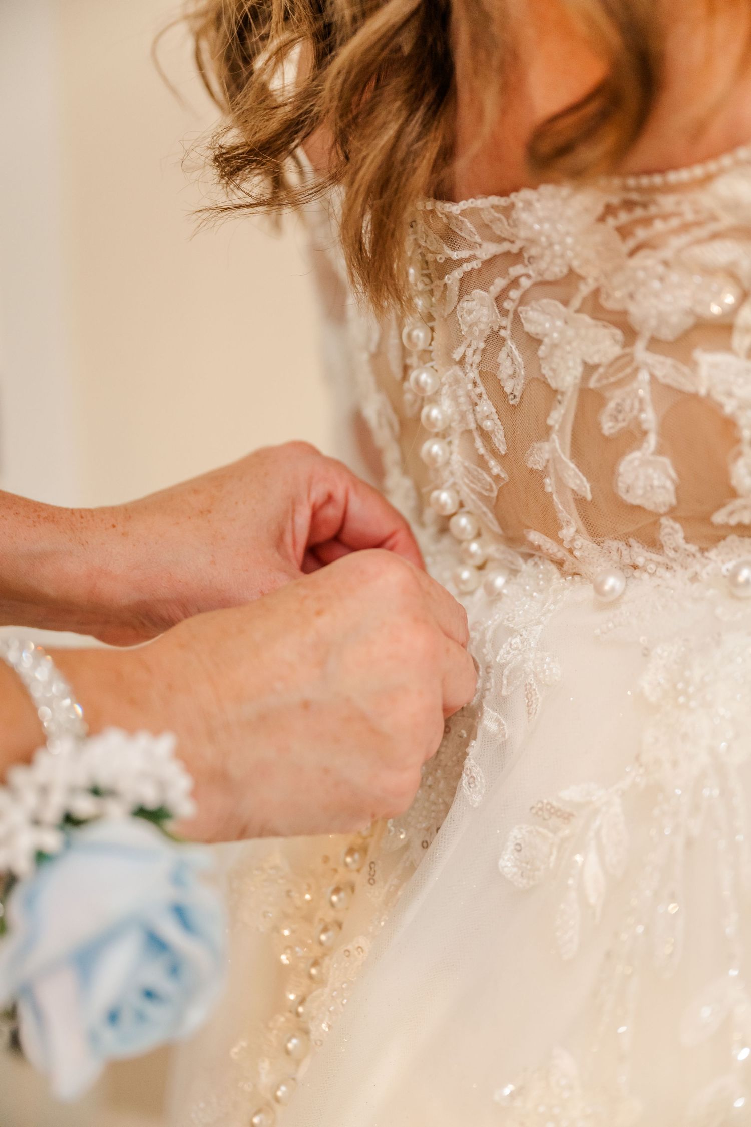 Close-up view of hands fastening delicate lace buttons on the back of an elegant white wedding dress.Four Seasons Barn Wedding & Event Venue