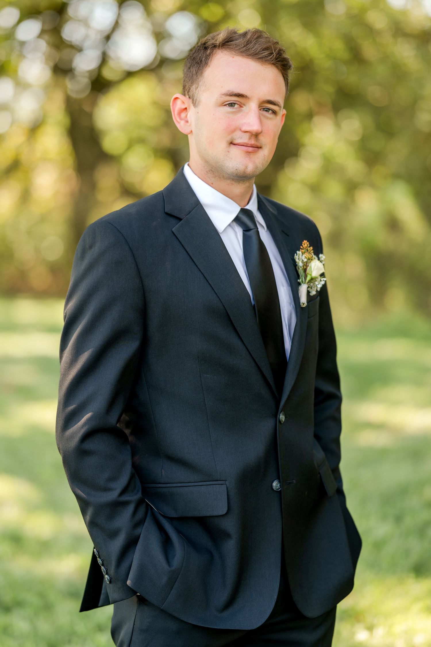Smiling groom in a navy suit and dark tie with a floral boutonniere stands with hands in pockets on a sunlit, tree-lined lawn.