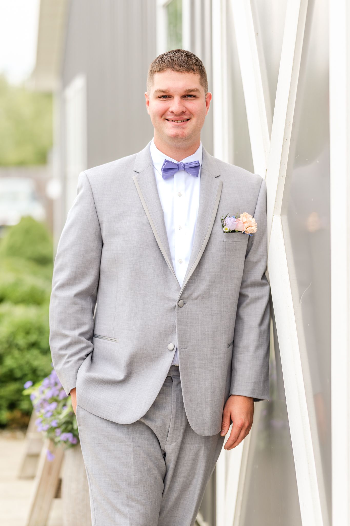 Smiling groom in a light gray suit, lavender bow tie and floral boutonniere stands beside a white building with greenery behind him.