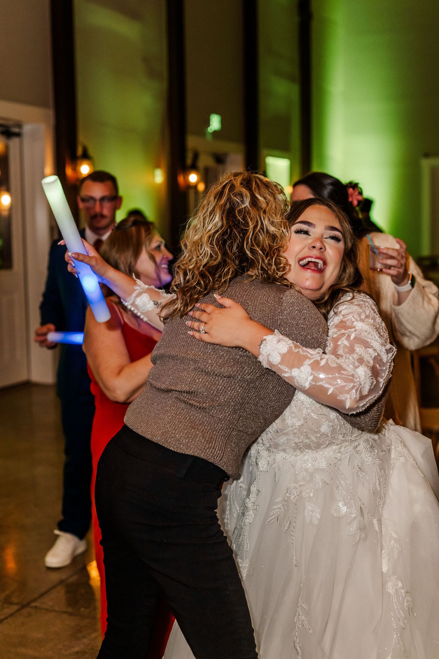 A bride in a white lace gown joyfully dances and embraces a guest at a wedding reception with green lighting.Four Seasons Barn Wedding