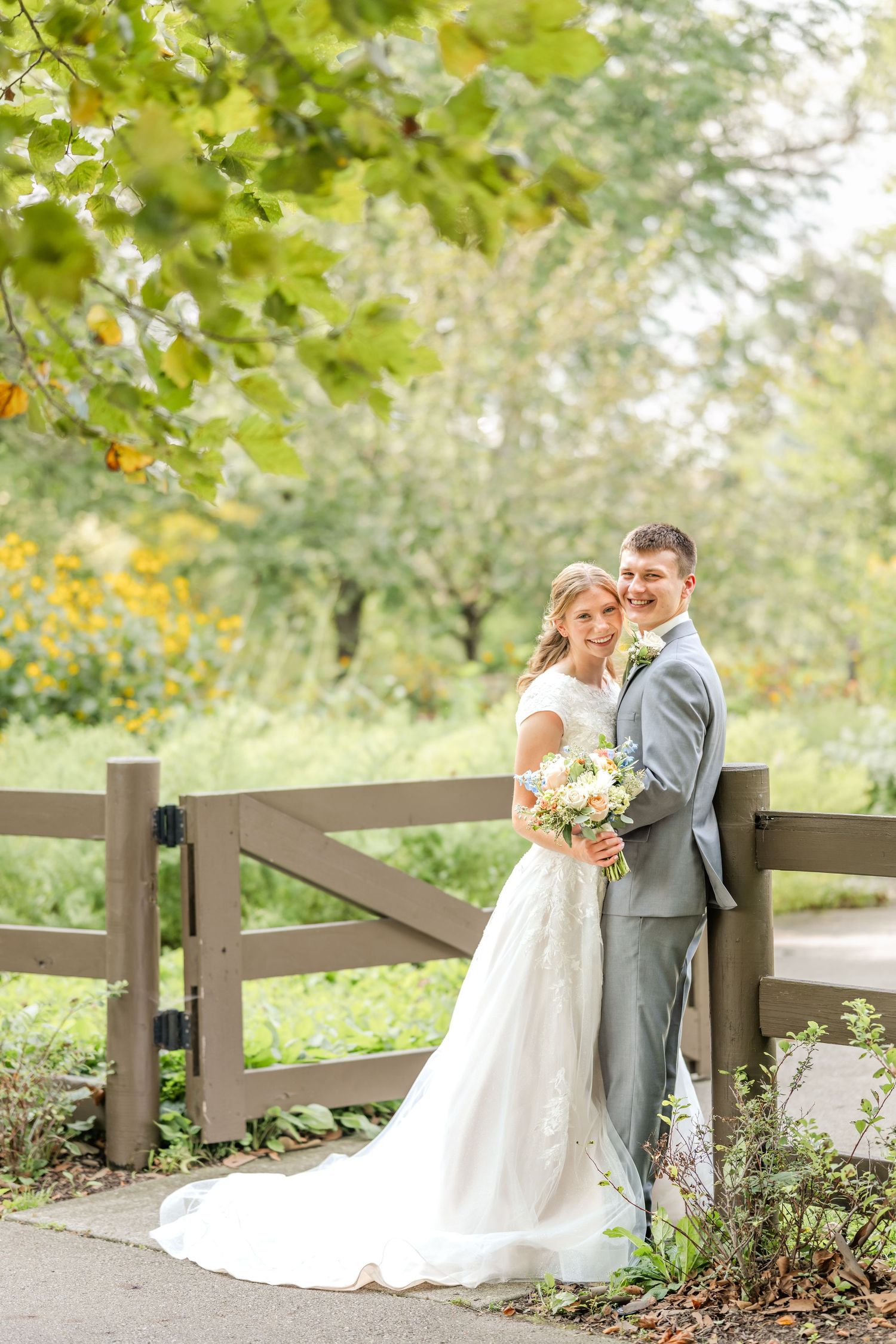 Bride in a lace wedding gown and groom in a gray suit embrace beside a wooden fence, holding a pastel bouquet in a sunny garden.