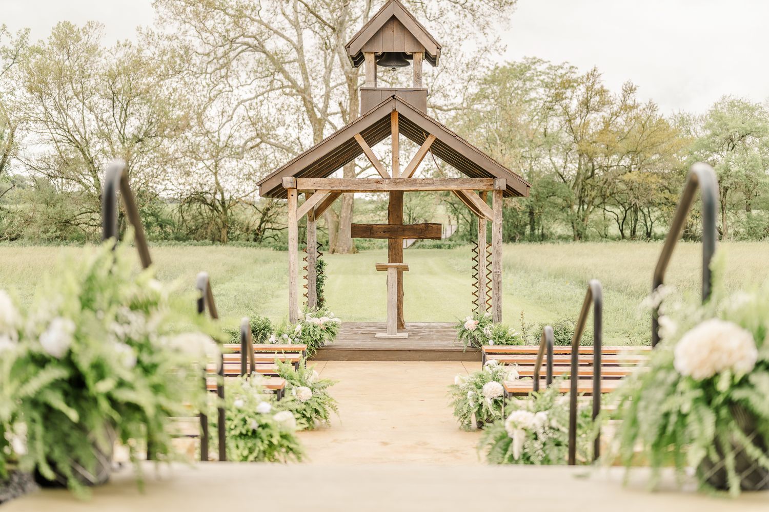 Outdoor wedding chapel with wooden cross altar, flanked by floral arrangements and pews on a lush green lawnThe Old Barns at Dry Run Farms.