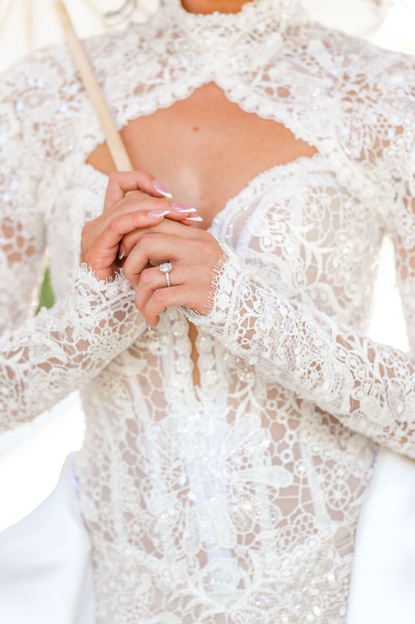 Bride in an ornate white lace long-sleeve wedding gown with keyhole neckline clasps her hands to show off a diamond engagement ring.